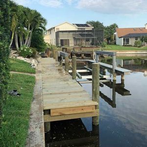 Wooden dock extends into a canal. A boat lift is attached. Green grass and houses are in the background.