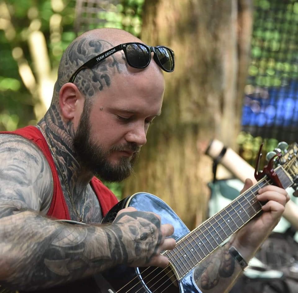 Man with tattoos plays guitar outdoors, wearing sunglasses.