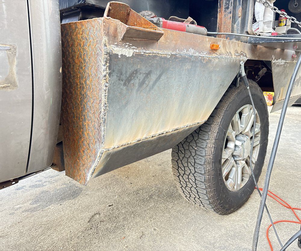Truck bed side constructed from welded metal, with tire and ground visible.