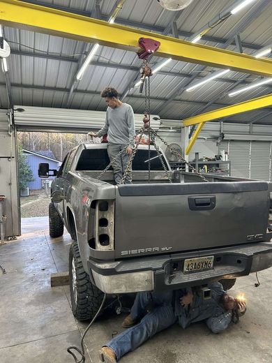 Two people working on a truck in a garage; one on the bed, one underneath. A crane is overhead.