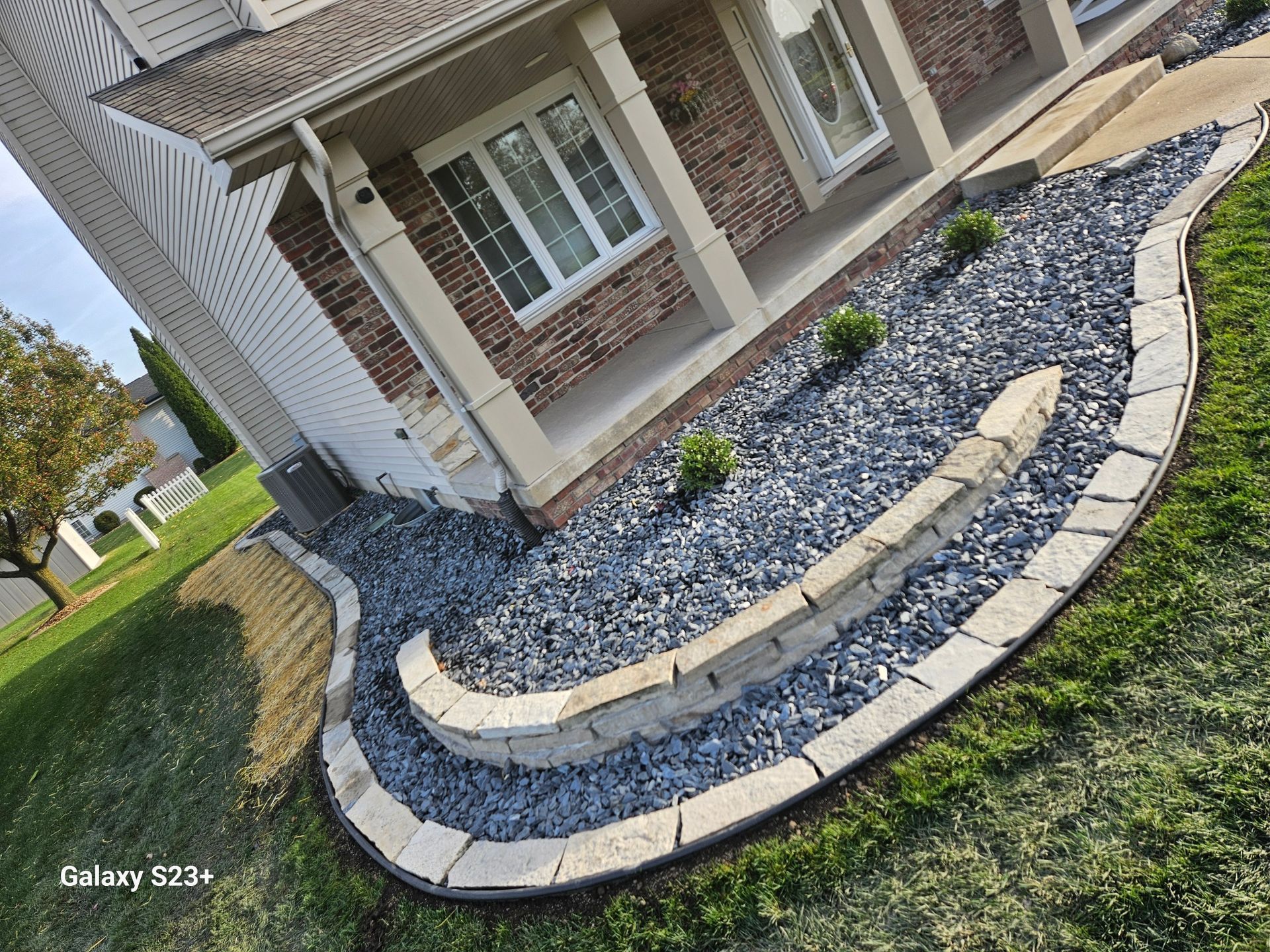A side view of a house foundation with a curved, tiered garden bed landscaped with dark grey gravel and stone edging.