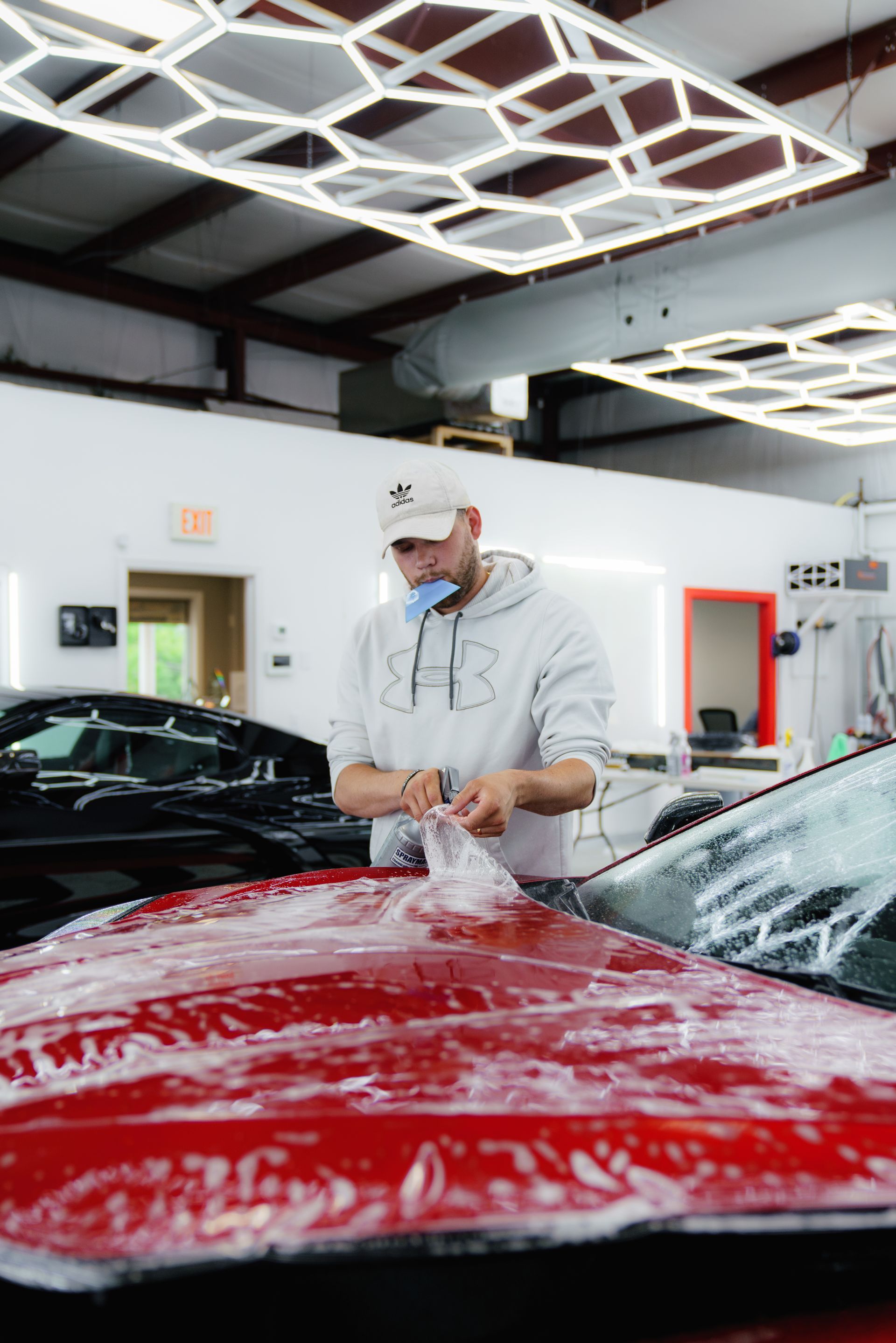 Man applying film to a red car's hood in a well-lit garage.