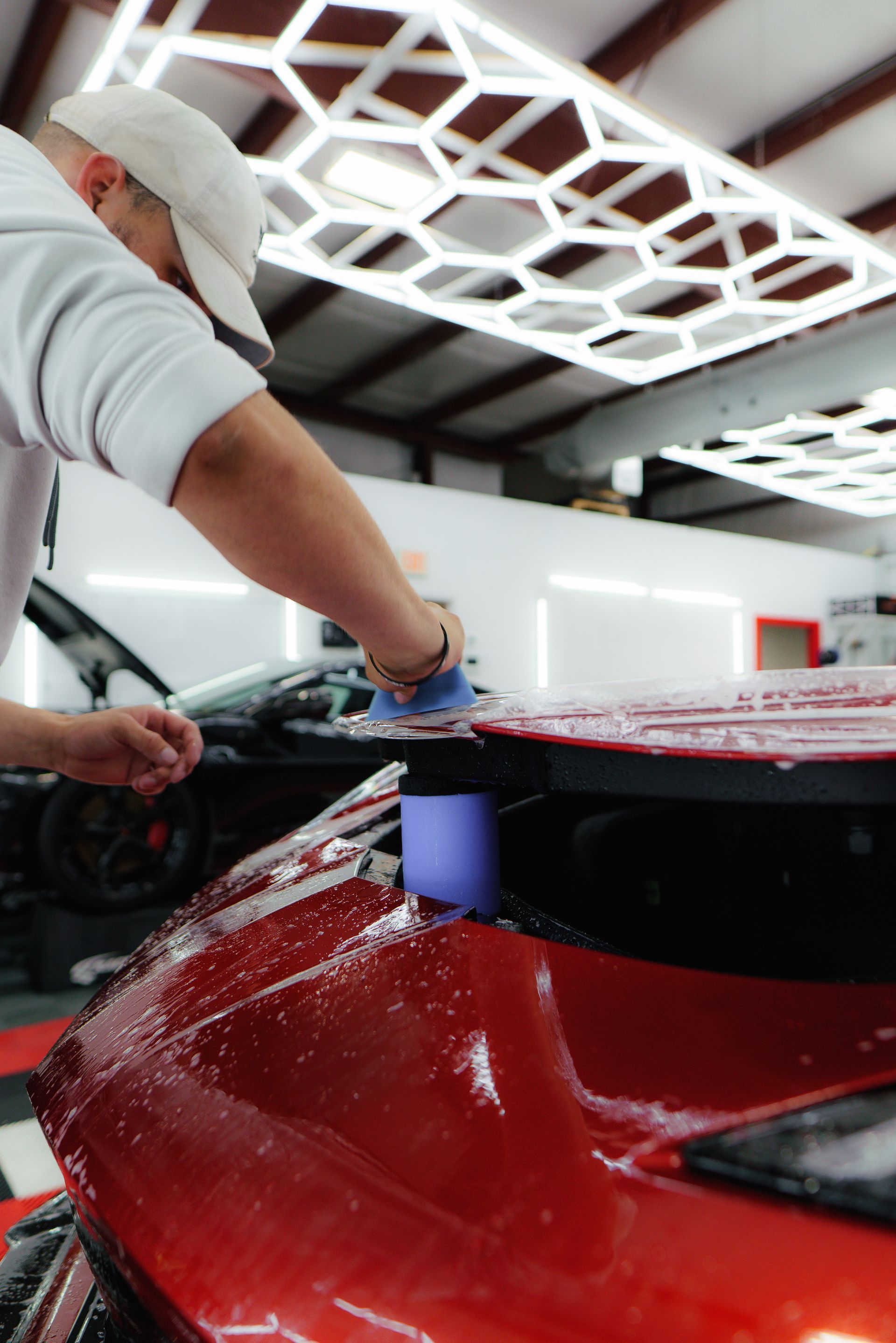 Man applies protective film to a red car hood in a shop, wearing a cap, under honeycomb lights.