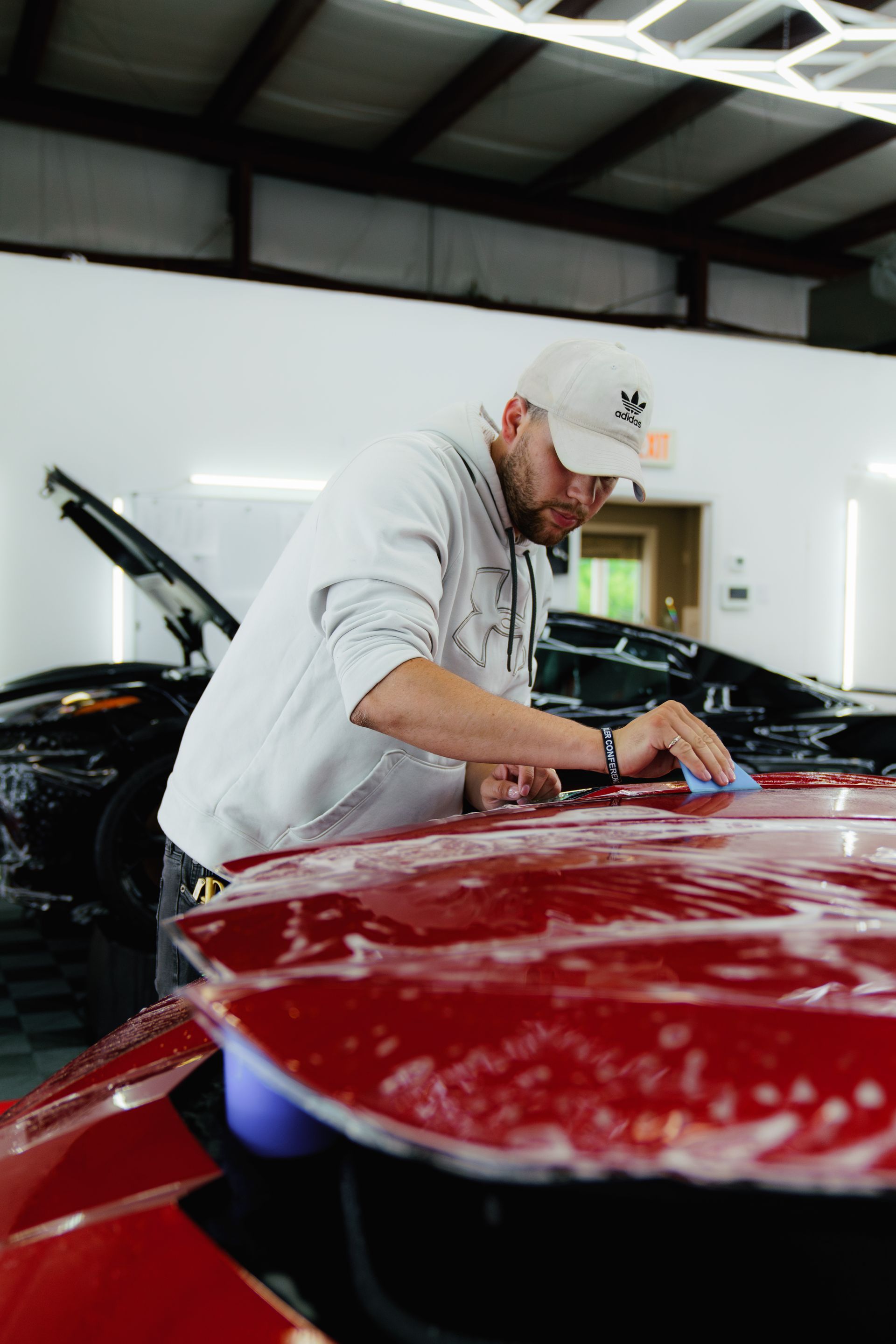 Man wearing a white cap, washes a red car's hood in a garage.