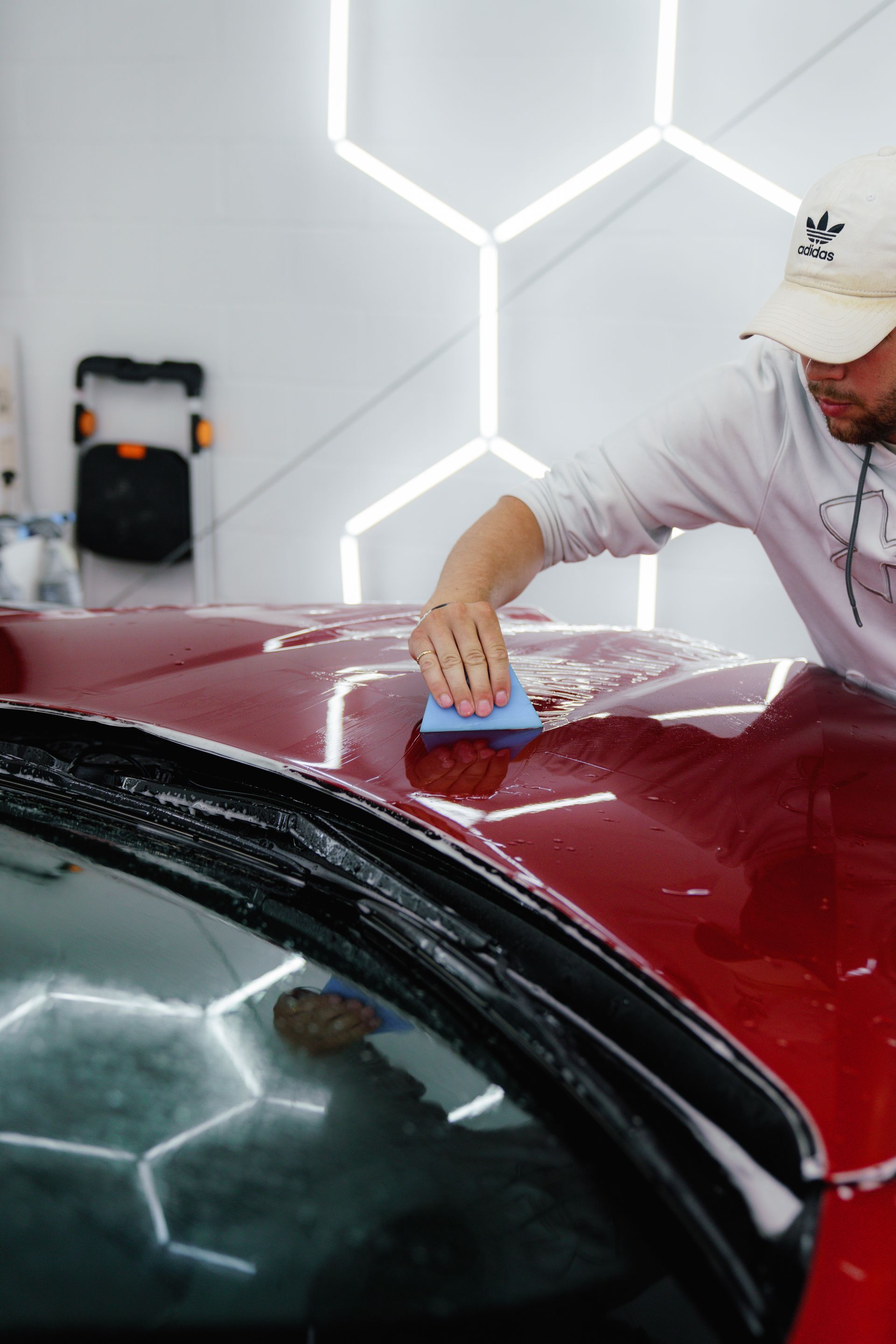 Person applying car wax to a red vehicle's hood, lit by hexagon-shaped lights.