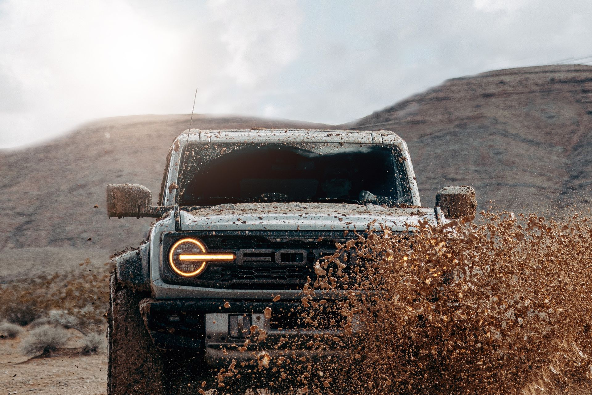 Ford Bronco driving through mud in a desert setting; front view, headlights on.