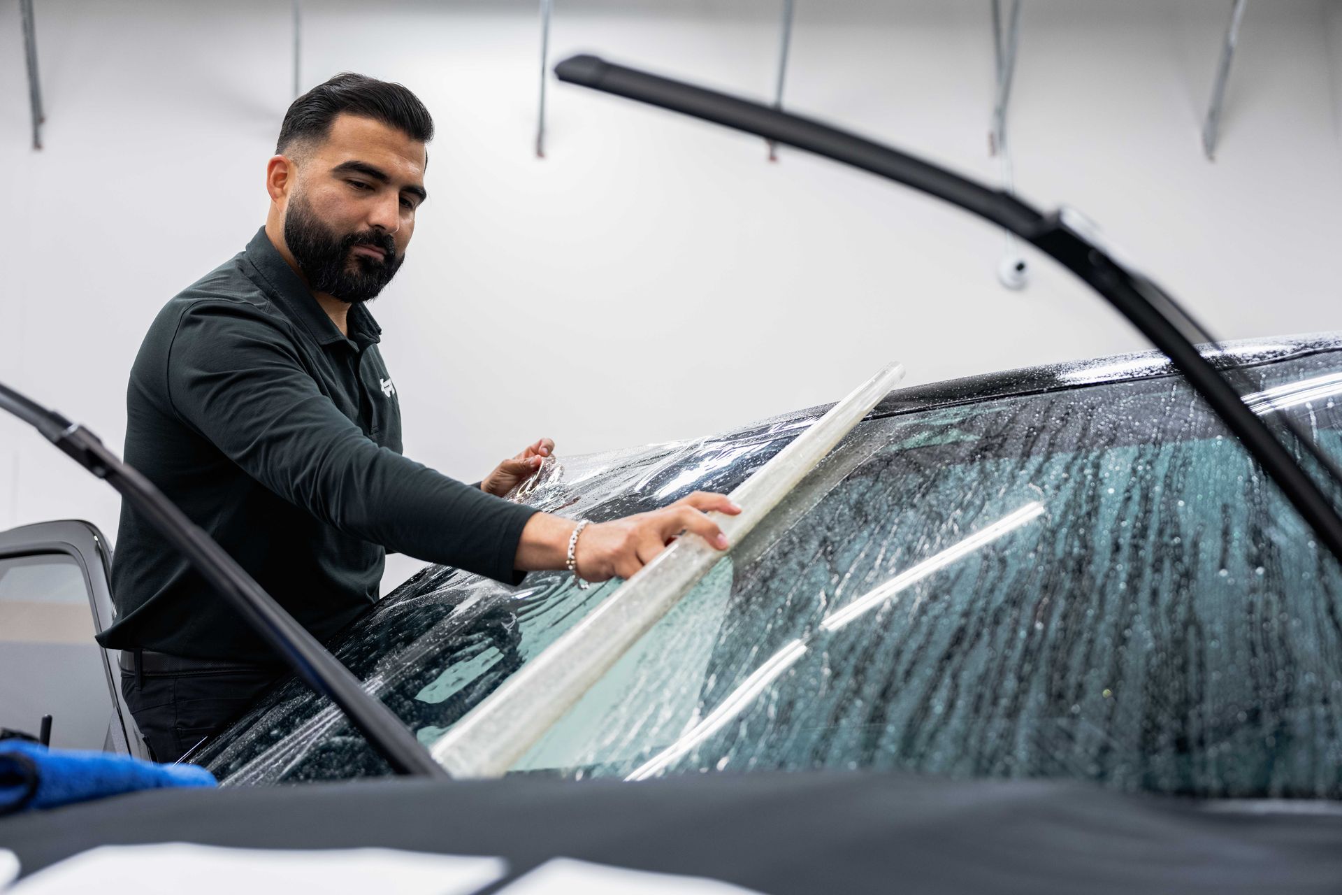 Man in dark shirt squeegeeing a wet car windshield in a bright garage.
