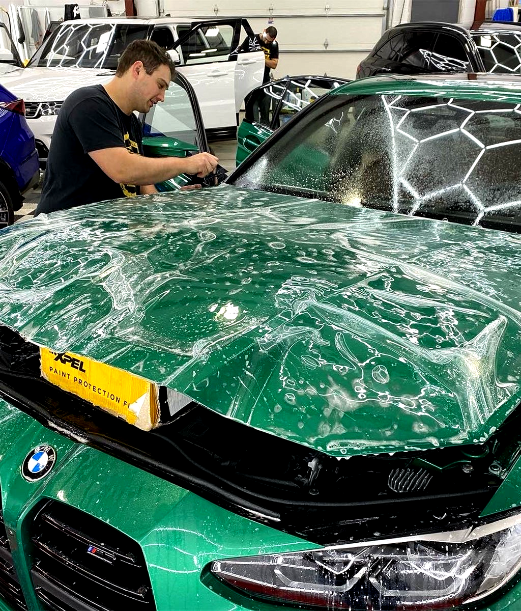 Man applying protective film to a green BMW car hood in a shop.