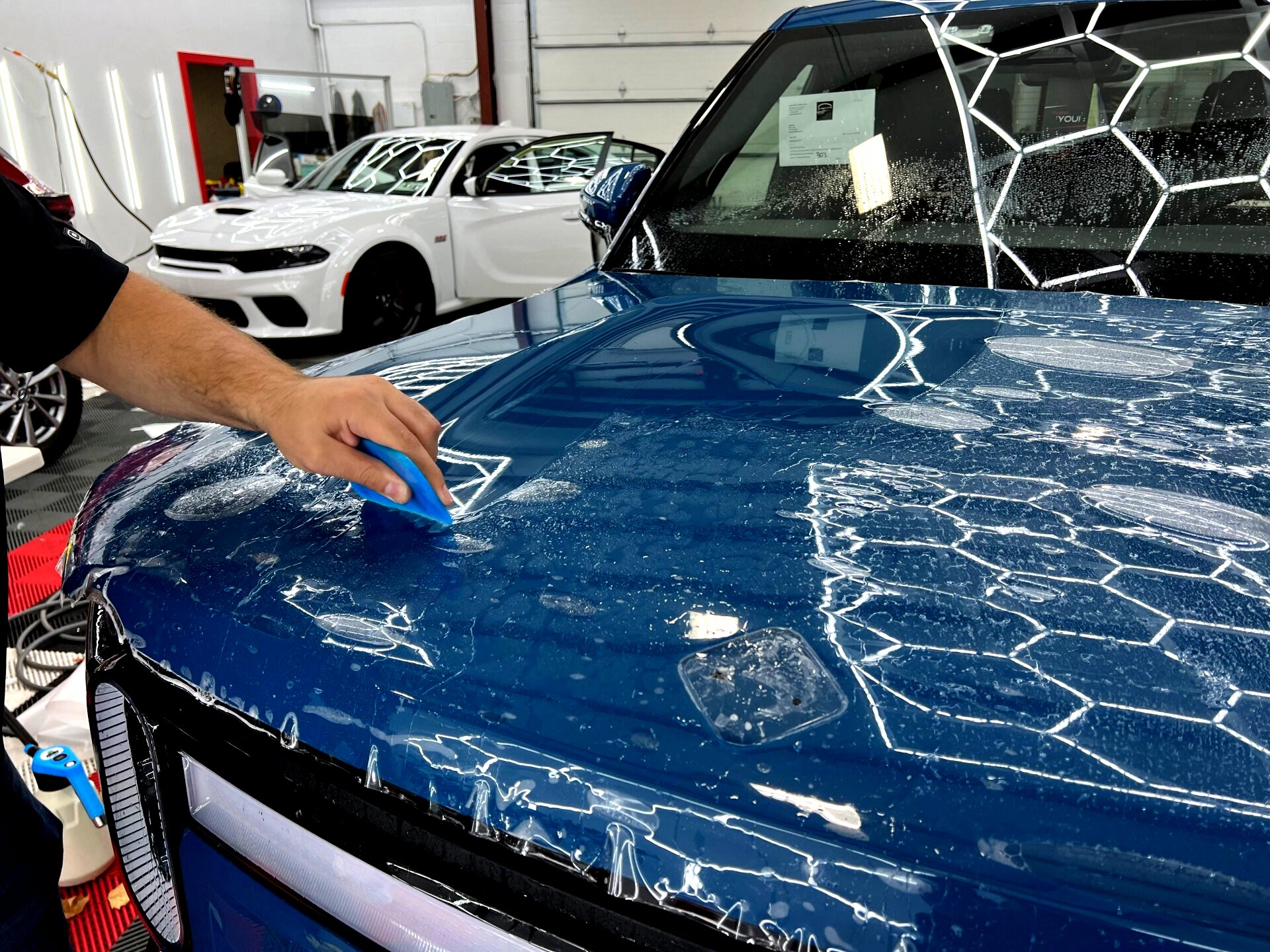 Person using a squeegee to apply protective film on a blue car hood in a garage. White car visible in the background.
