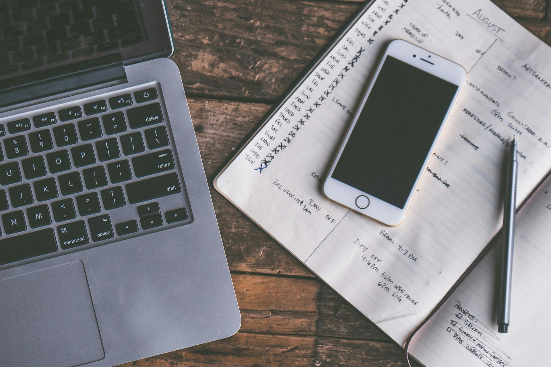 Laptop, smartphone, and pen on handwritten notes on a wooden desk.