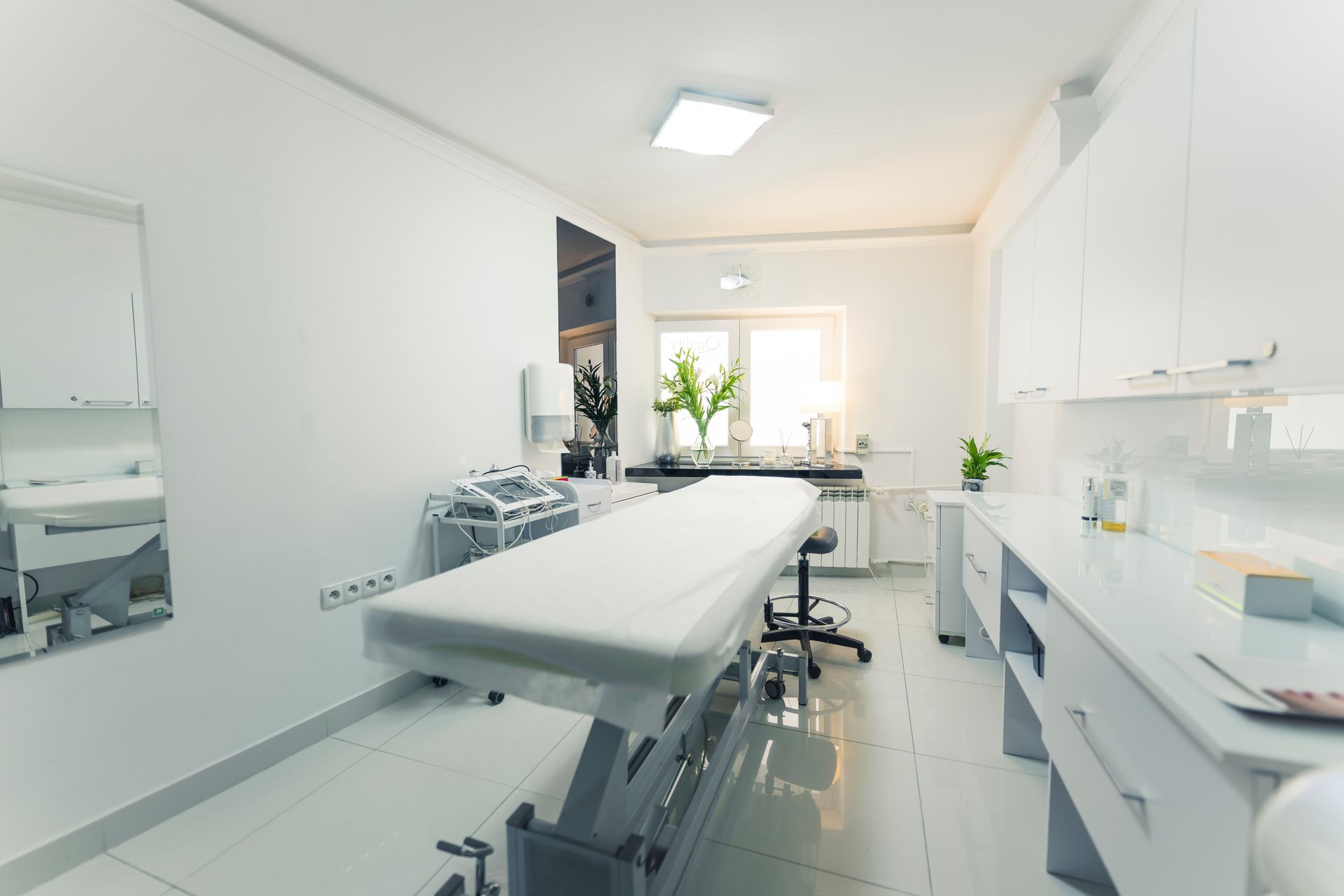 Bright, modern medical treatment room with an exam table, white cabinets, and a window with plants.