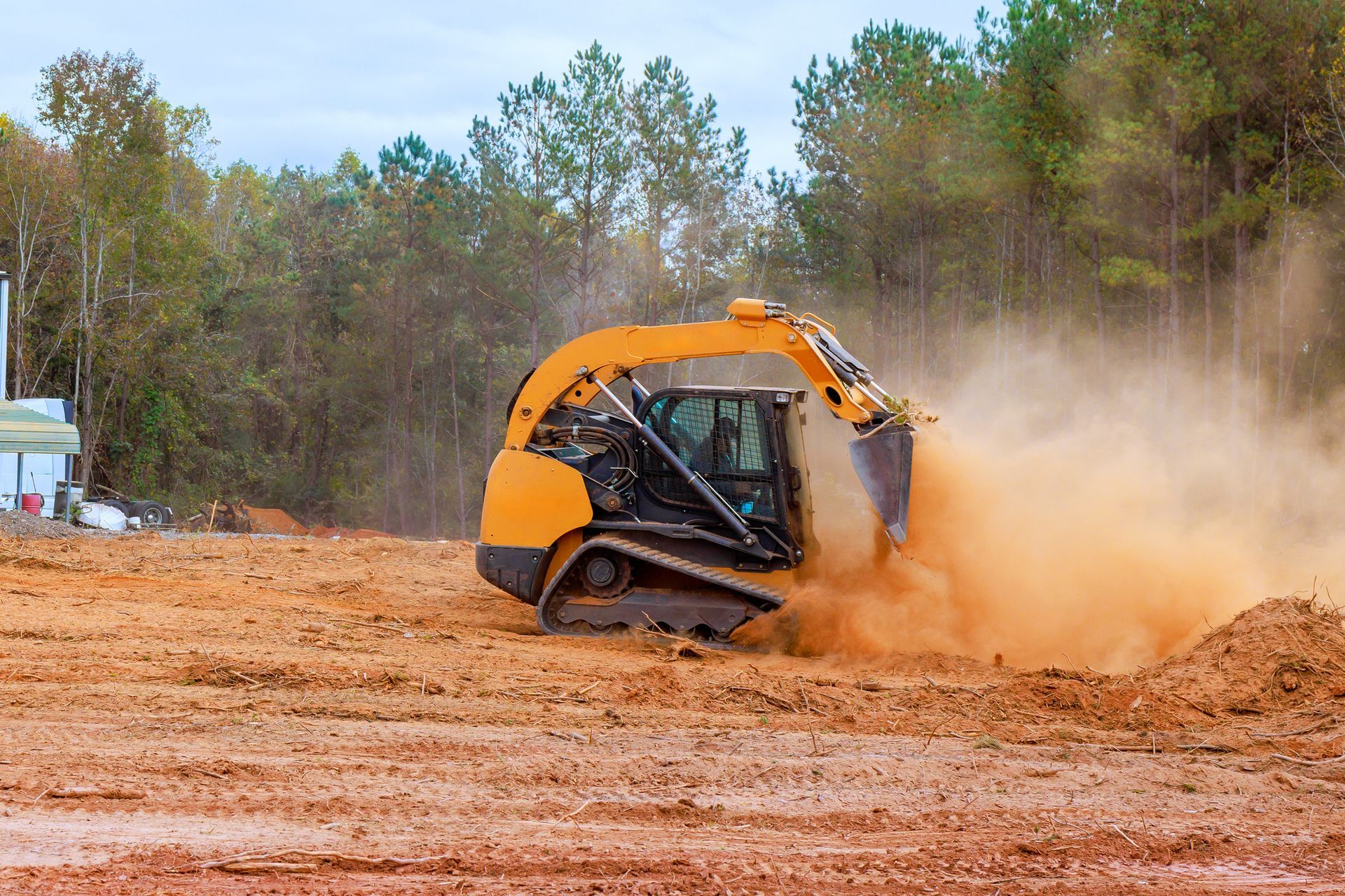 Yellow skid steer on tracks, kicking up dust while working on a construction site with trees in the background. Yellow skid steer on tracks, kicking up dust while working on a construction site with trees in the background.