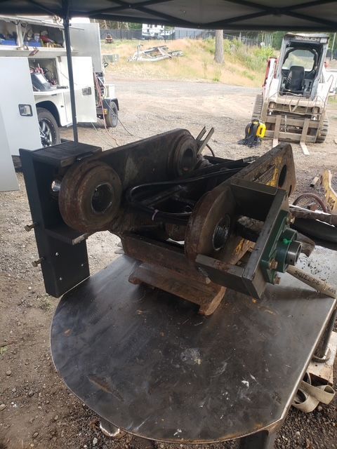 Heavy machinery components on a table outdoors, with a white vehicle and skid steer in the background.