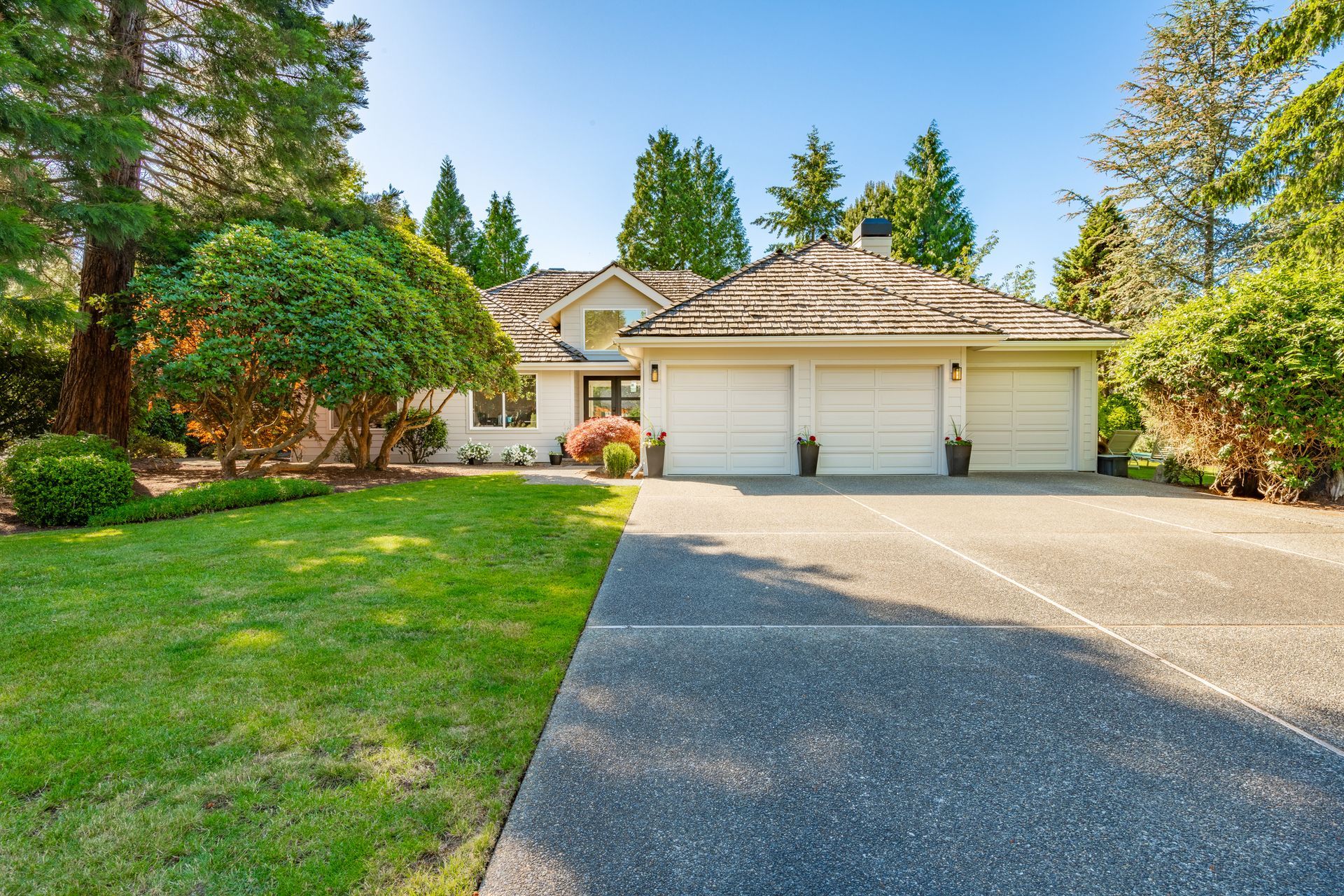 Beige house with three-car garage, concrete driveway, and lush green lawn, surrounded by trees under a bright sky.