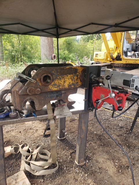 Yellow excavator part being worked on, held on a table. A red drill is attached. Outdoors.
