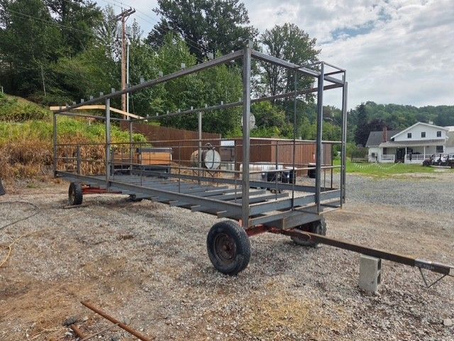 Steel frame trailer with a cage-like structure, sitting on a gravel lot near trees and buildings.