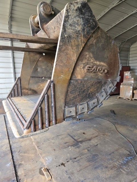 Large, rusty excavator bucket in a metal building. The bucket has teeth and 
