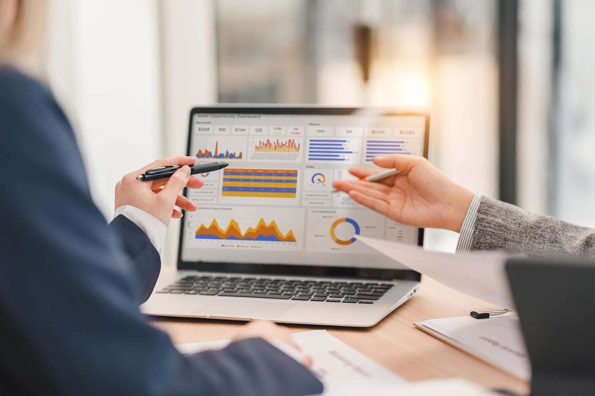 Two people analyzing financial charts on a laptop, pointing at data; indoor office setting.