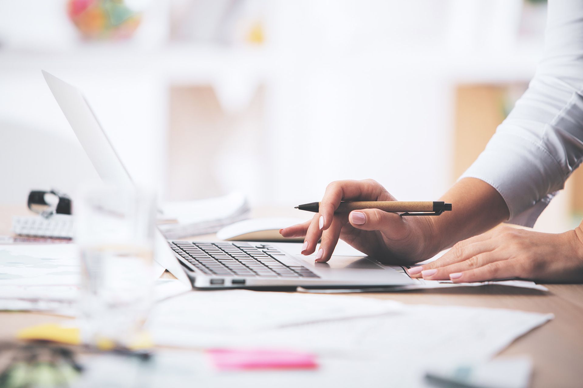 Hands typing on laptop, holding pen, working at desk with papers.