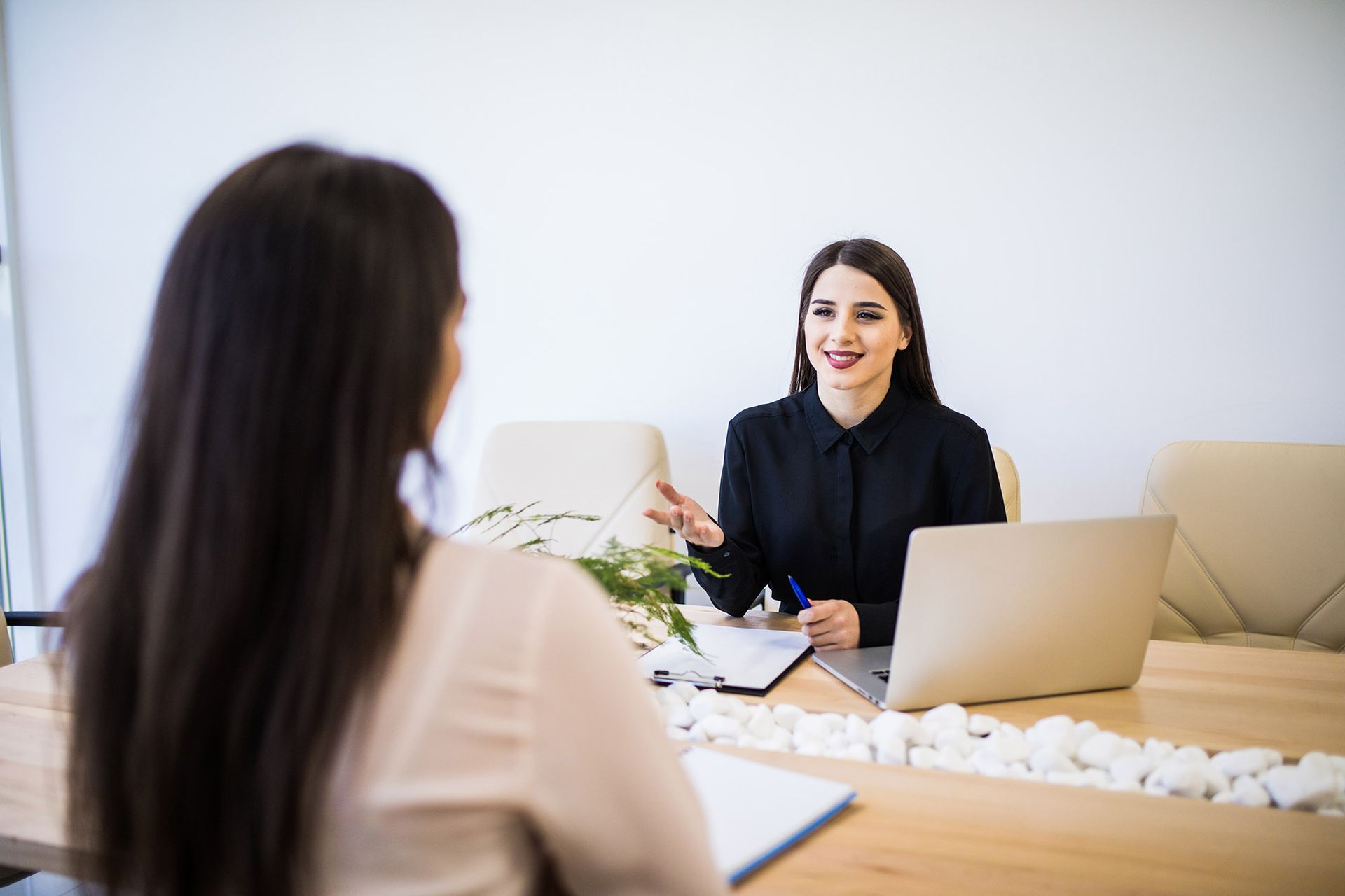 Woman in black suit interviewing another woman at a table with a laptop and paperwork.
