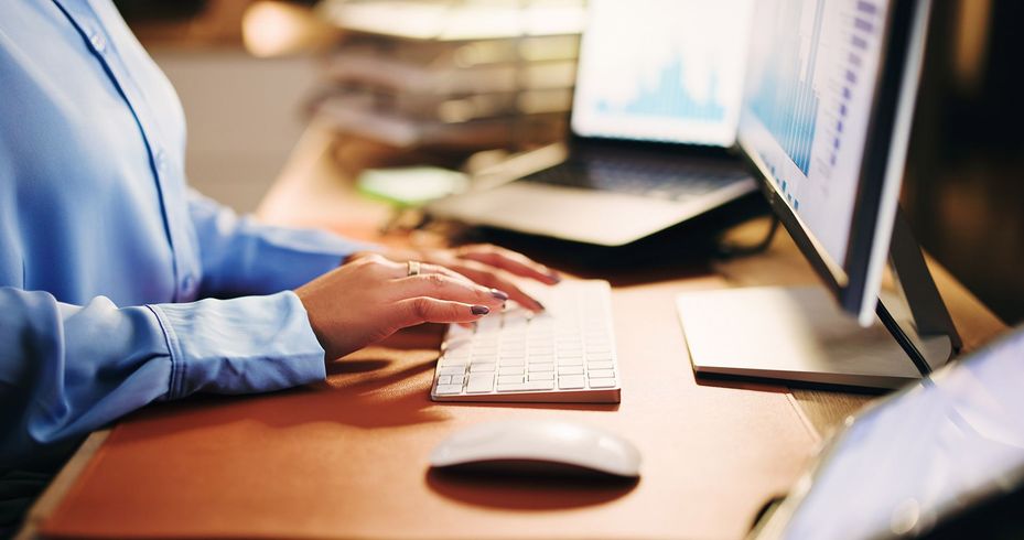 Person typing on a keyboard at a desk with a monitor and laptop displaying charts.