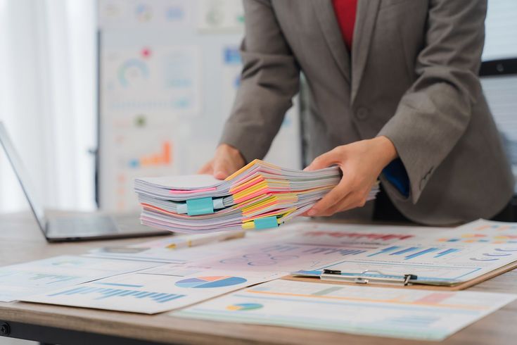 Person in suit holding stack of documents with colorful tabs, near a laptop and charts.