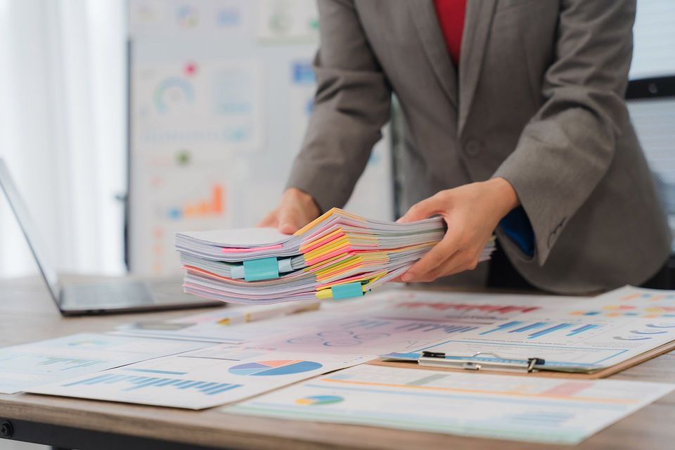 Person in suit holding stack of documents with colorful tabs, near a laptop and charts.