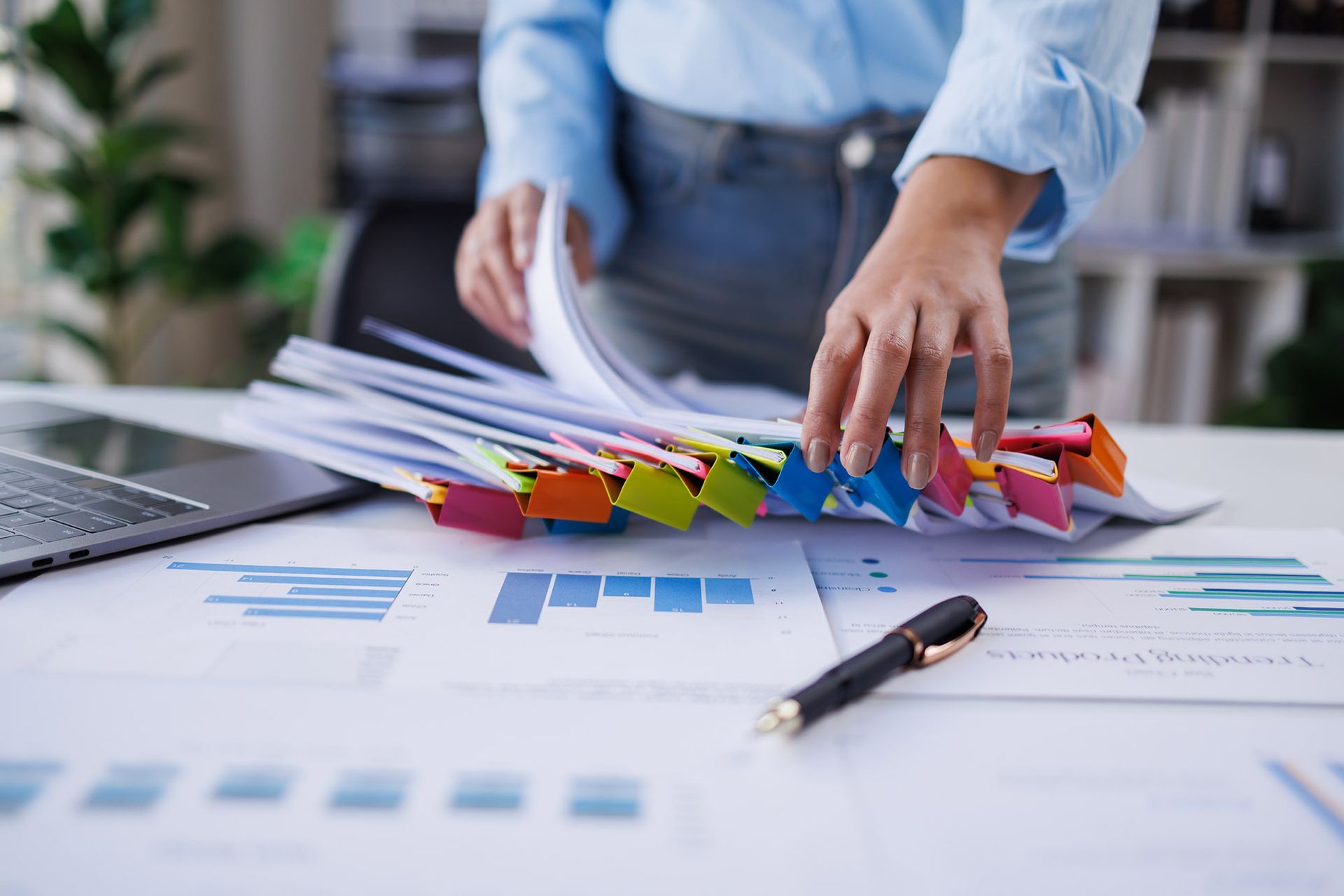 Woman organizing a stack of papers with colorful binder clips on a desk with a laptop and pen.