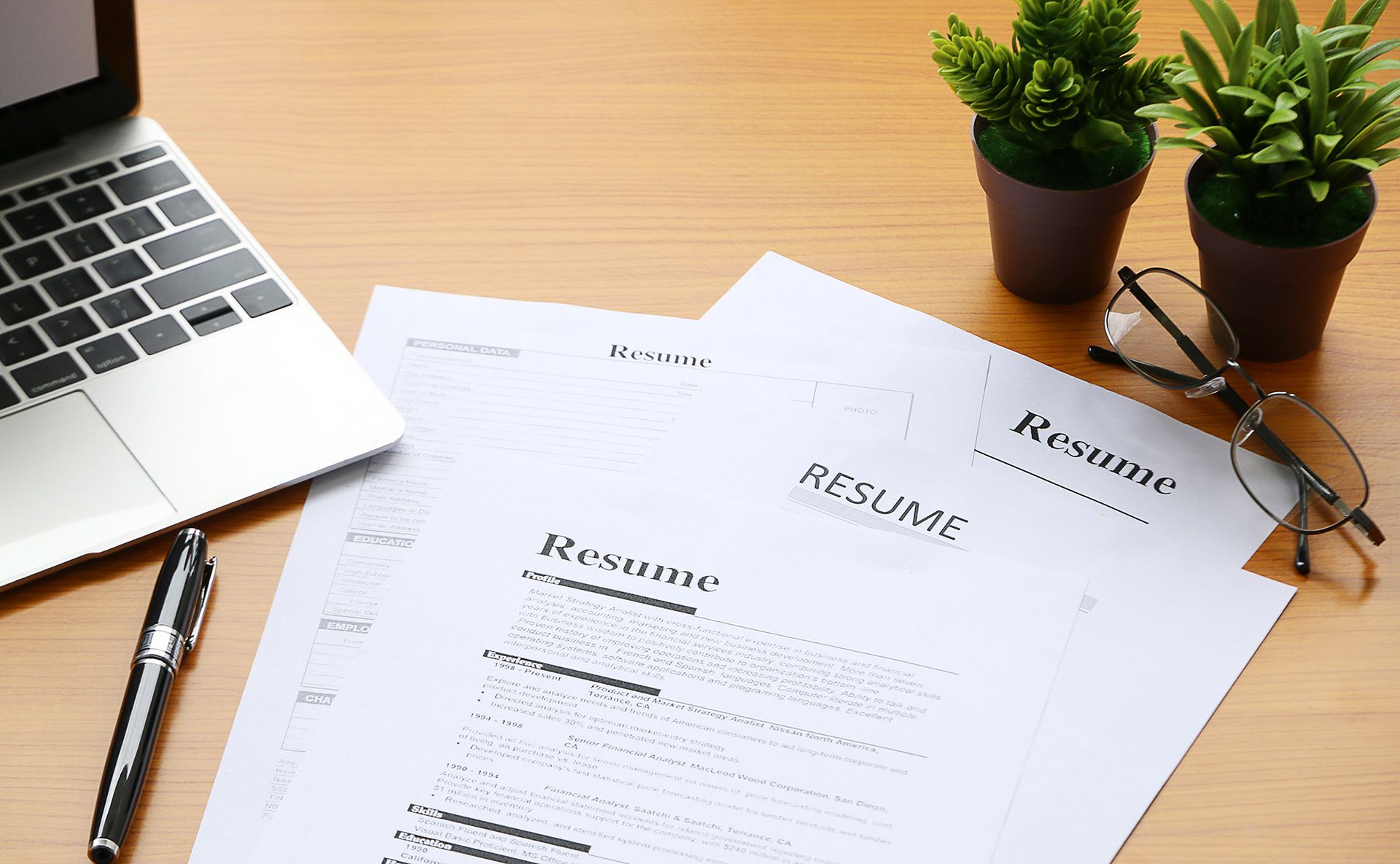 Laptop, resume, pen, and glasses on a wooden desk with small potted plants.