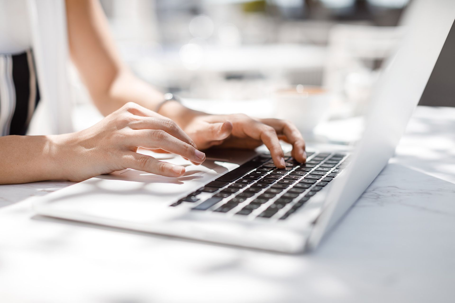 Woman's hands typing on a laptop computer outdoors at a white table in sunlight.