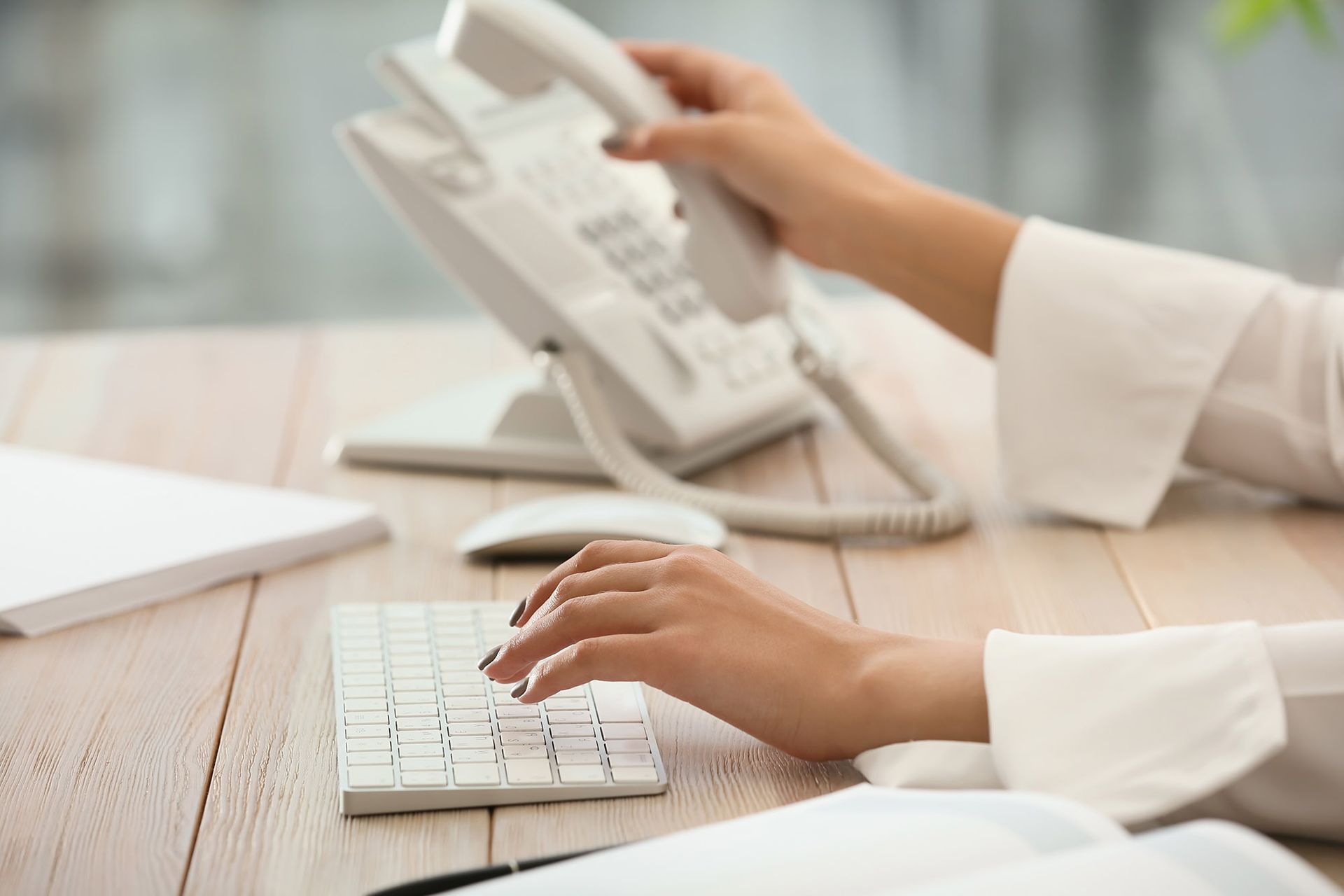 Person using a keyboard while holding a phone receiver in an office setting.
