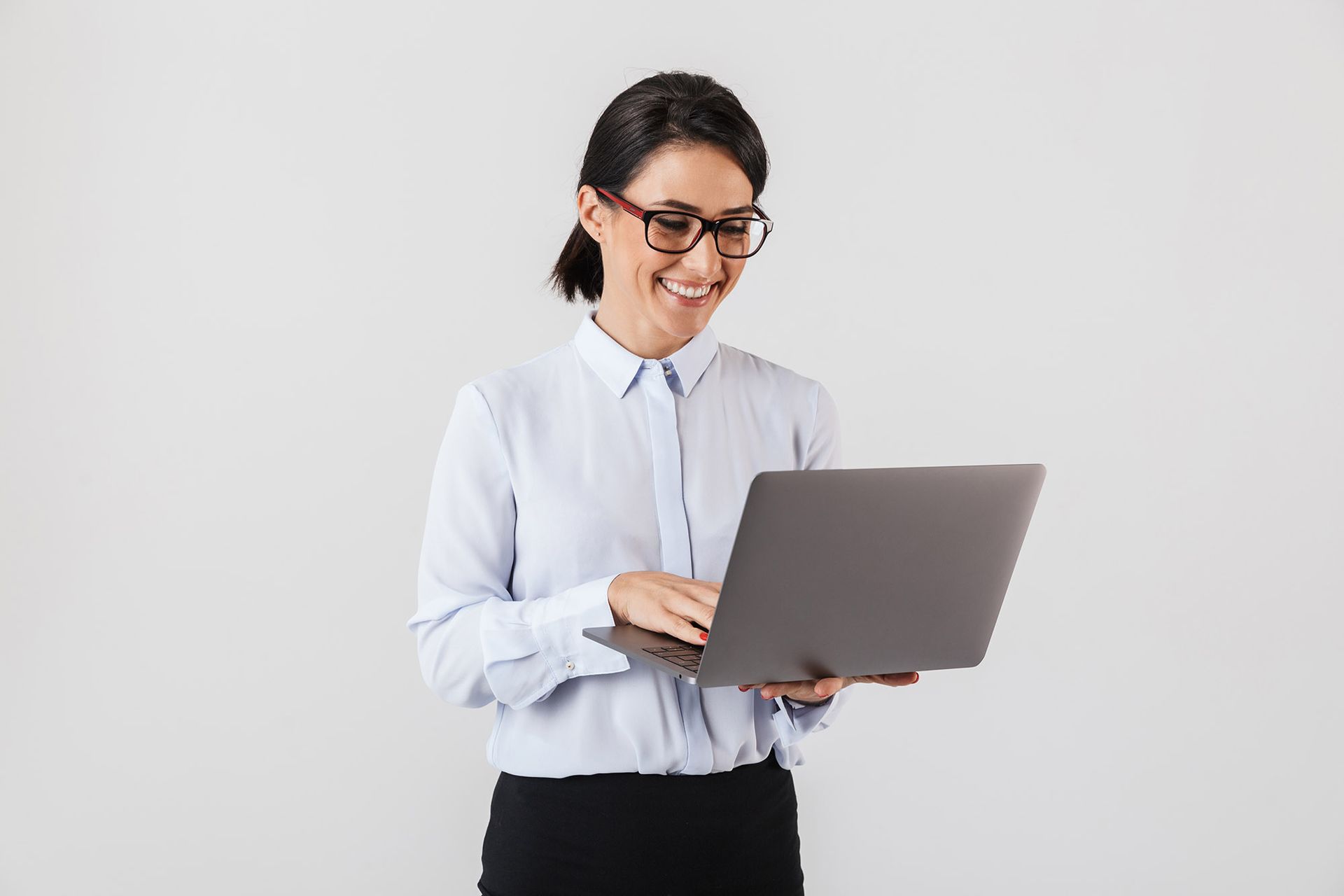 Woman in glasses smiles while typing on a laptop. Against a white background, she wears a blue button-down and black skirt.