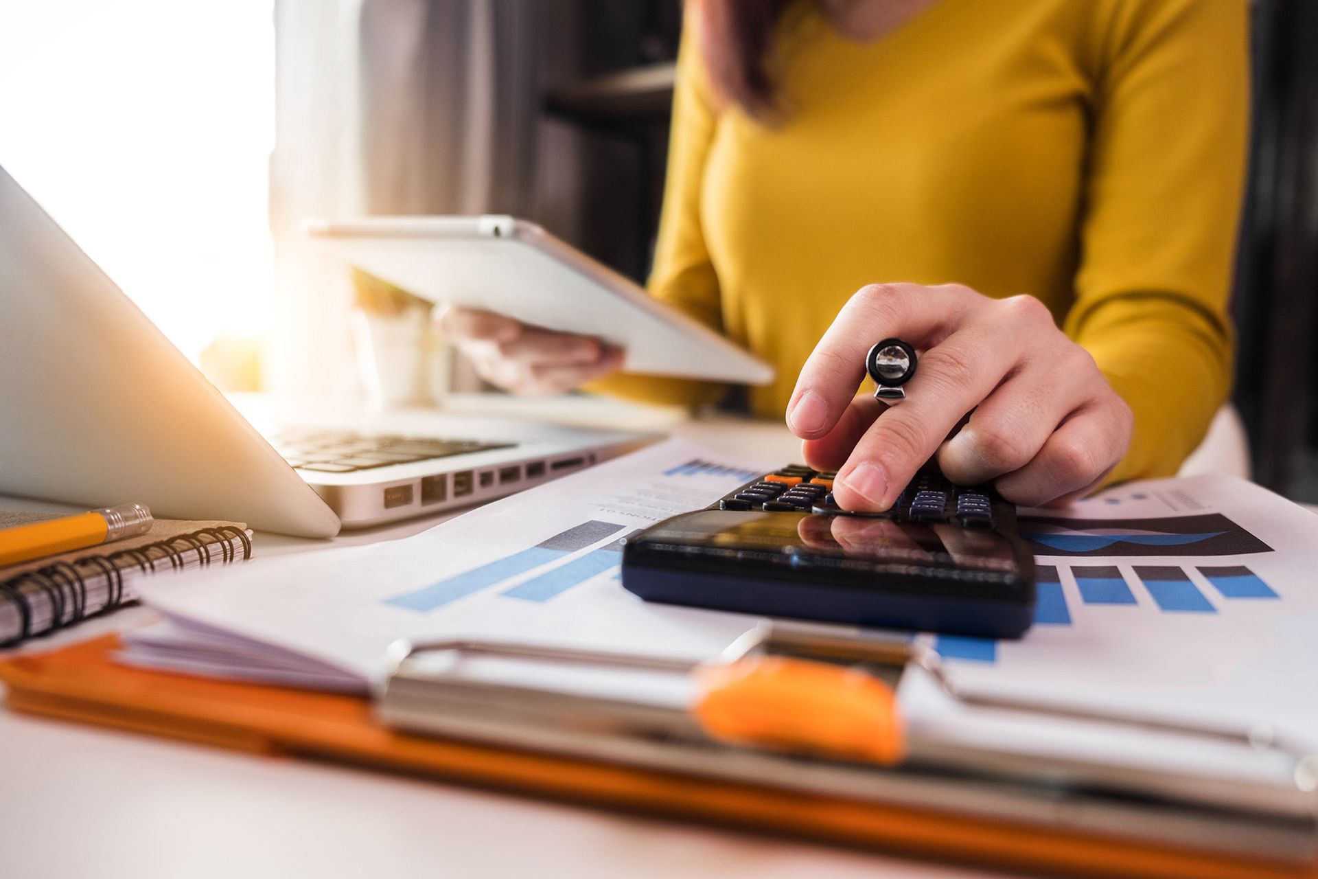 Woman in yellow sweater using a calculator, with laptop, tablet, and financial documents on a desk.