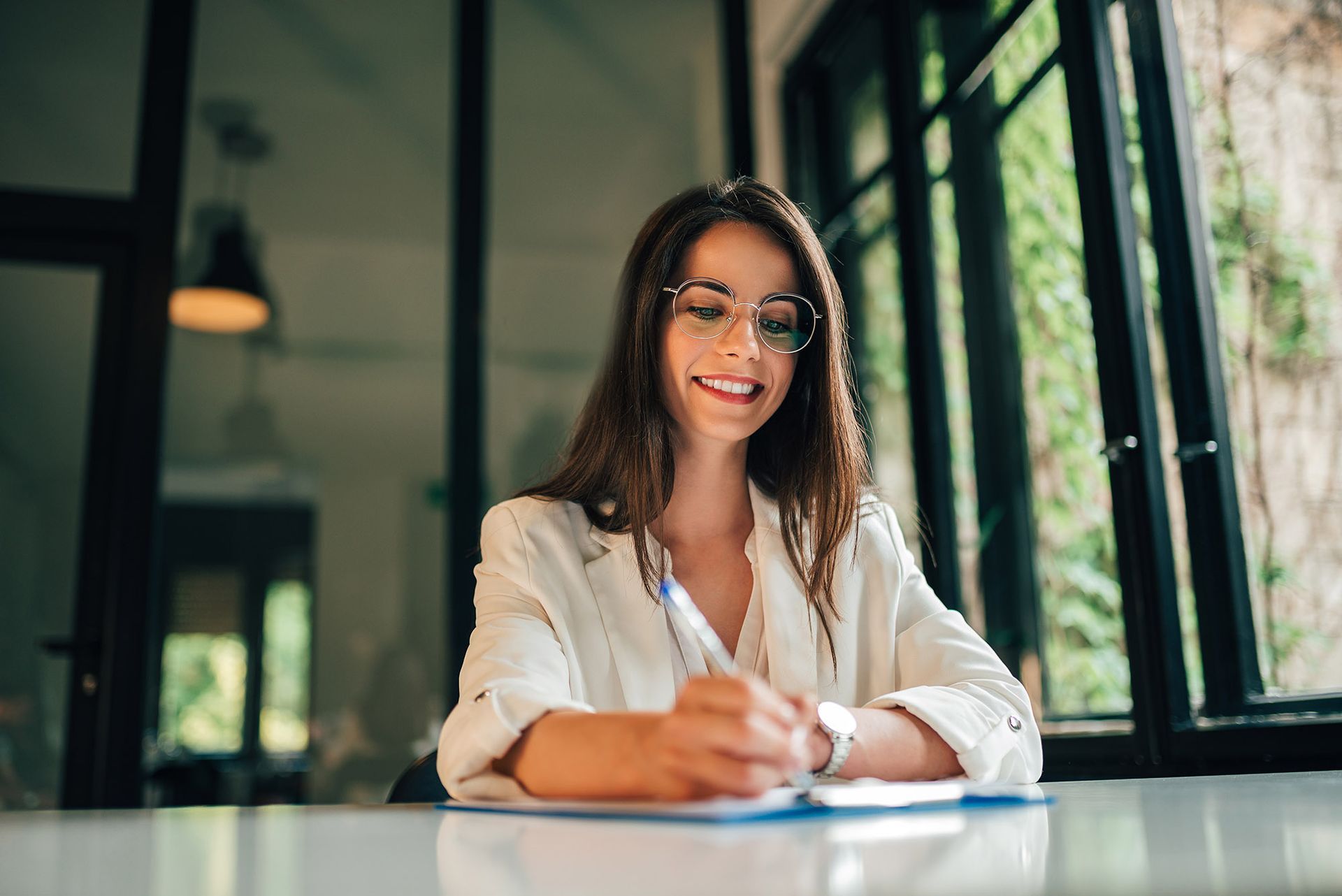 Woman in glasses writing at a table, smiling, with a window and plants in the background.