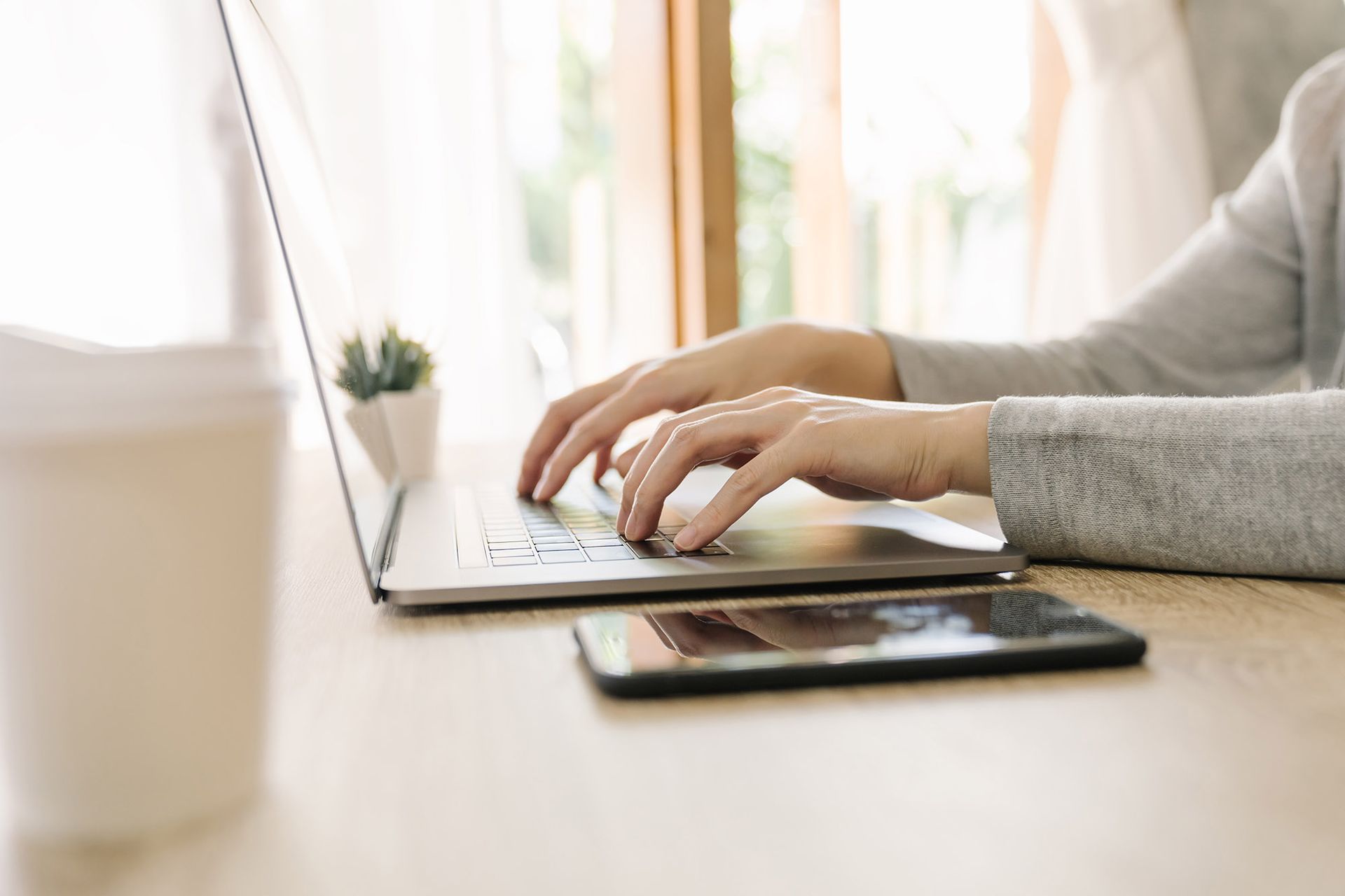Person typing on a laptop at a wooden desk with a phone, a coffee cup, and a small plant.