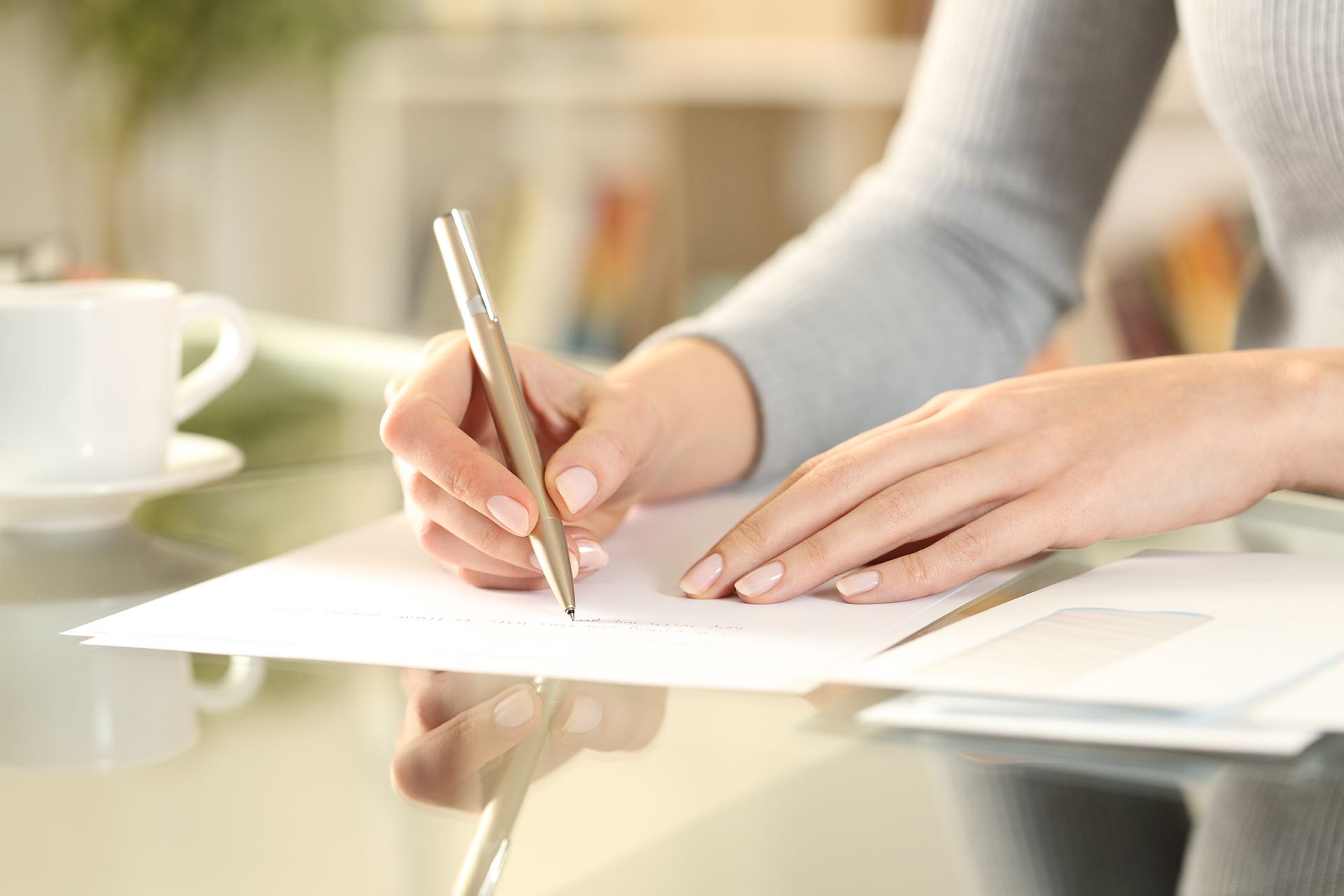 Woman writing on paper with a pen at a desk, near a cup and envelopes.