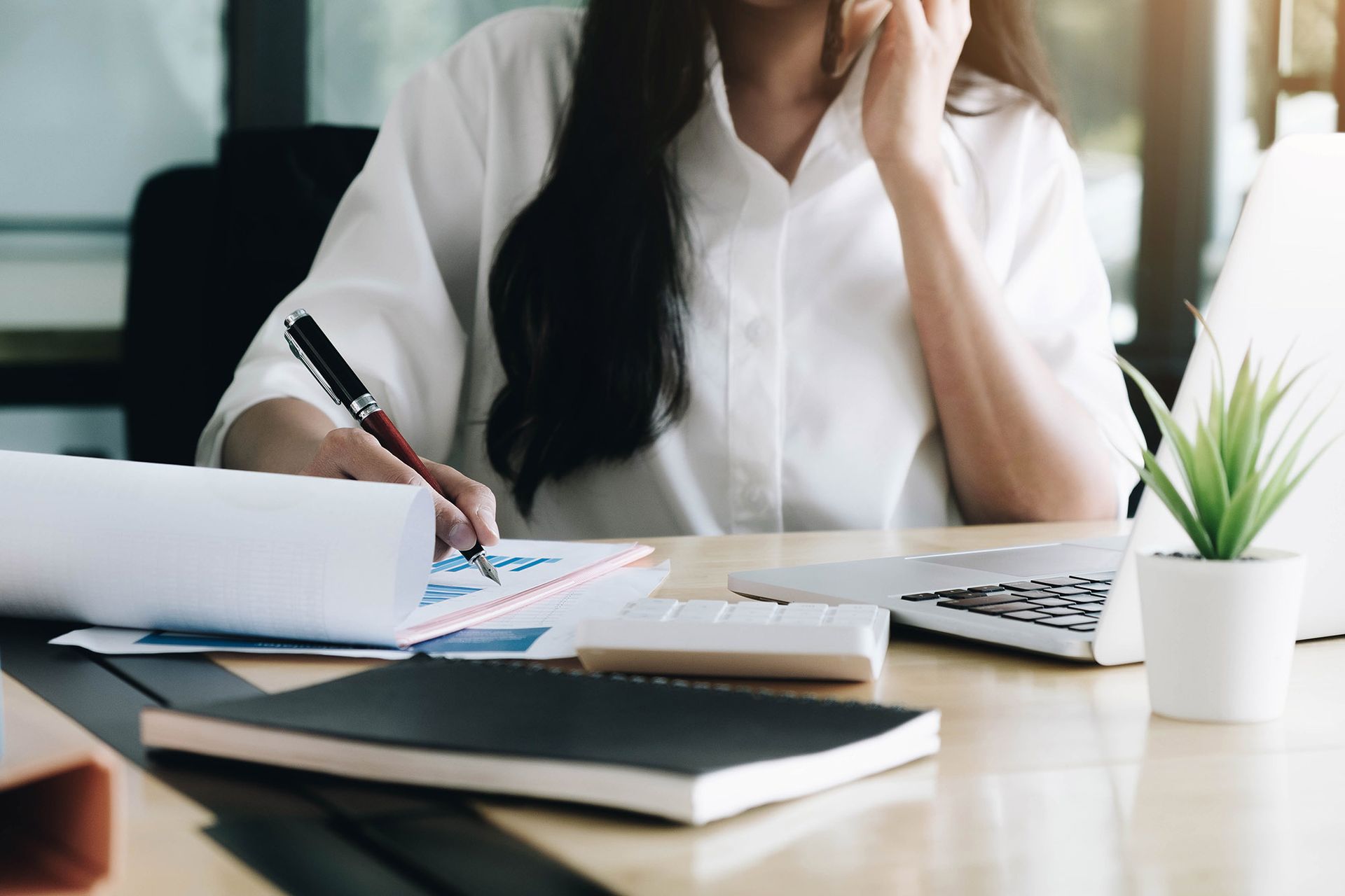 Woman in white shirt writing at a desk with papers, laptop, calculator, and plant.