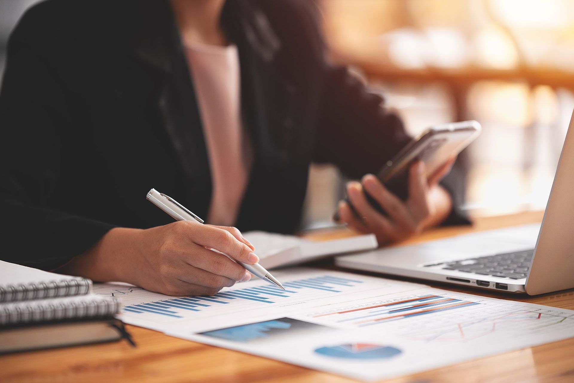 Woman in black blazer analyzes charts with a pen, holding a phone, laptop and notepad on desk.
