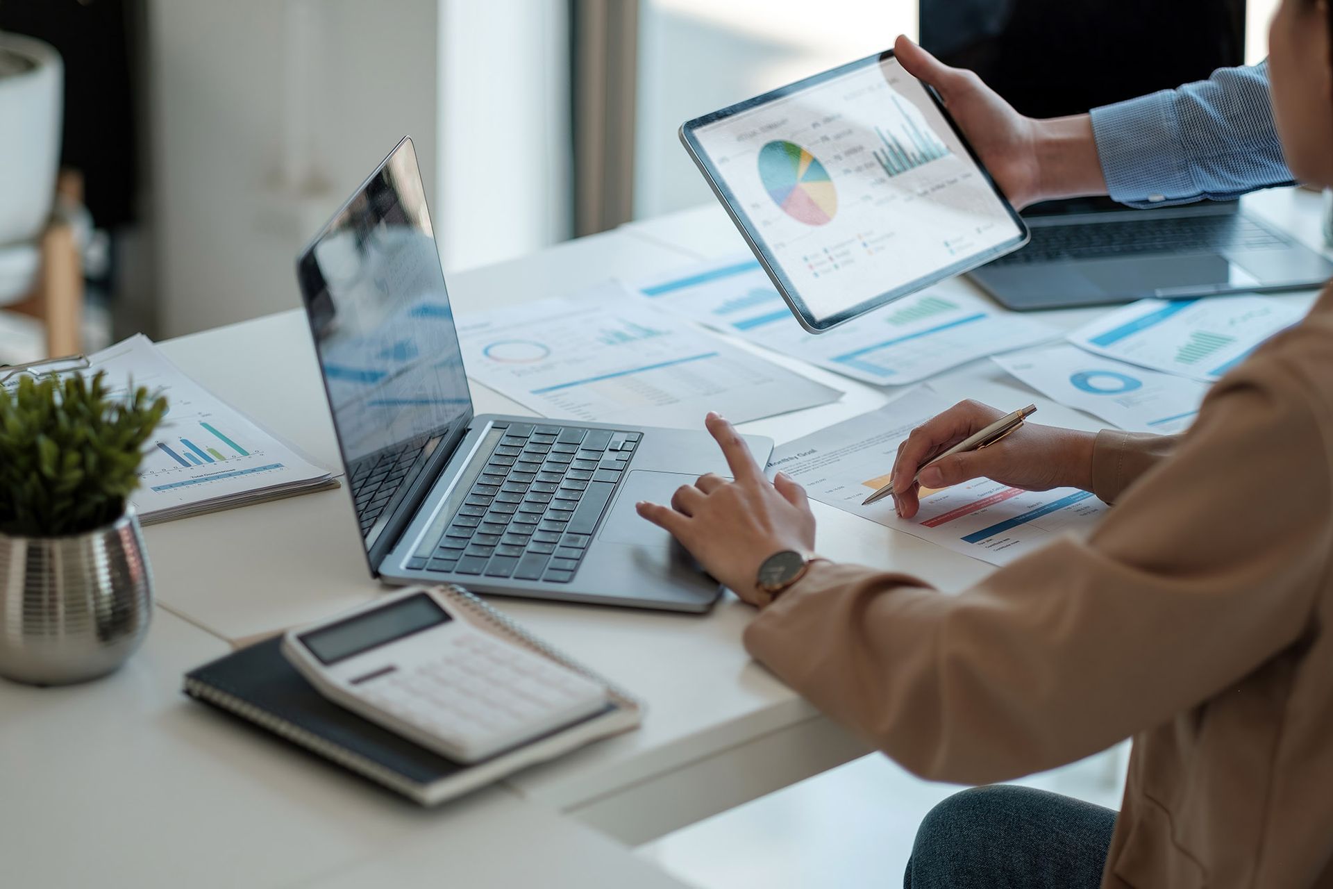Two people review financial data on a laptop, tablet, and printed papers at a desk.