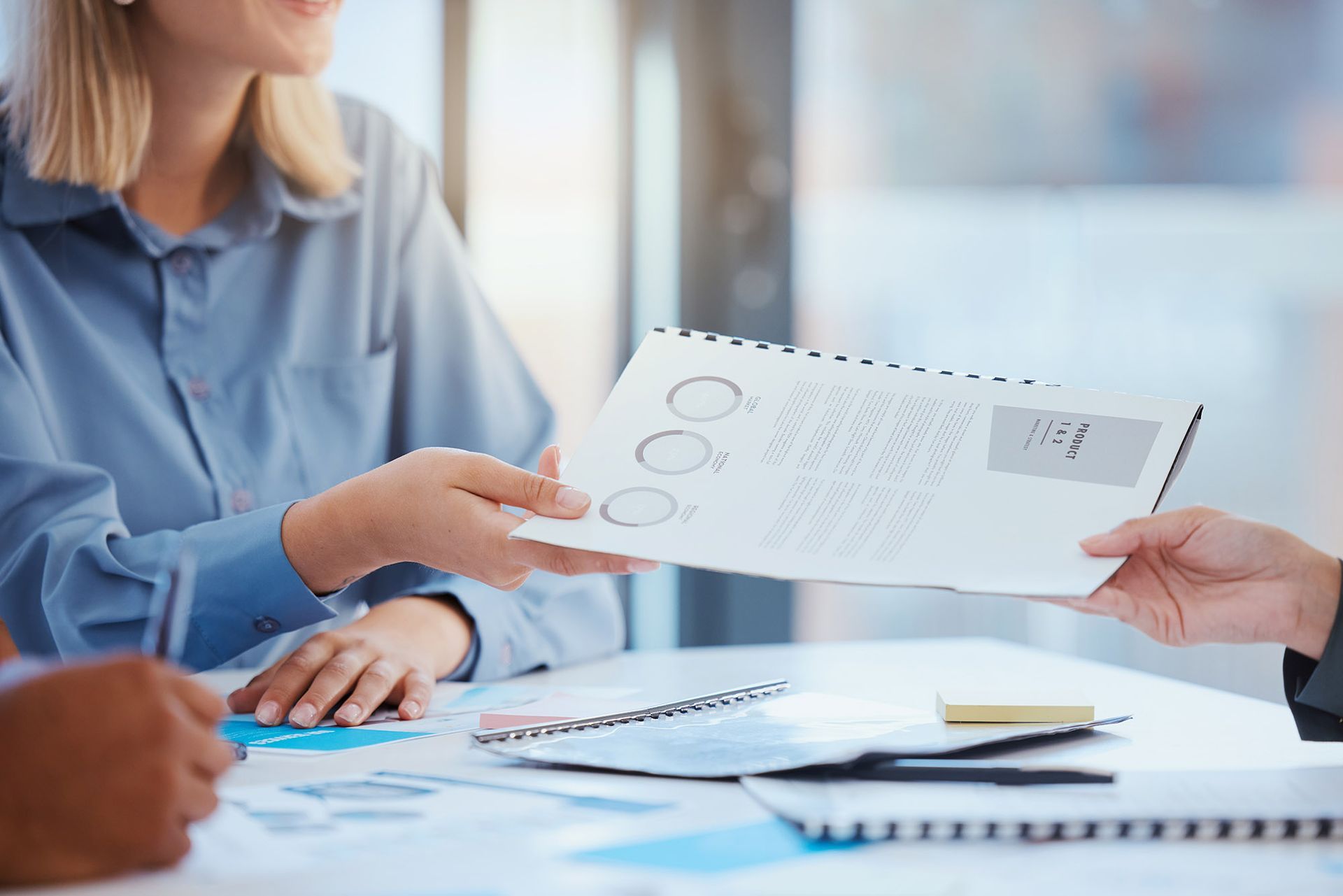 Person handing document with charts to another person at a table, likely in an office.
