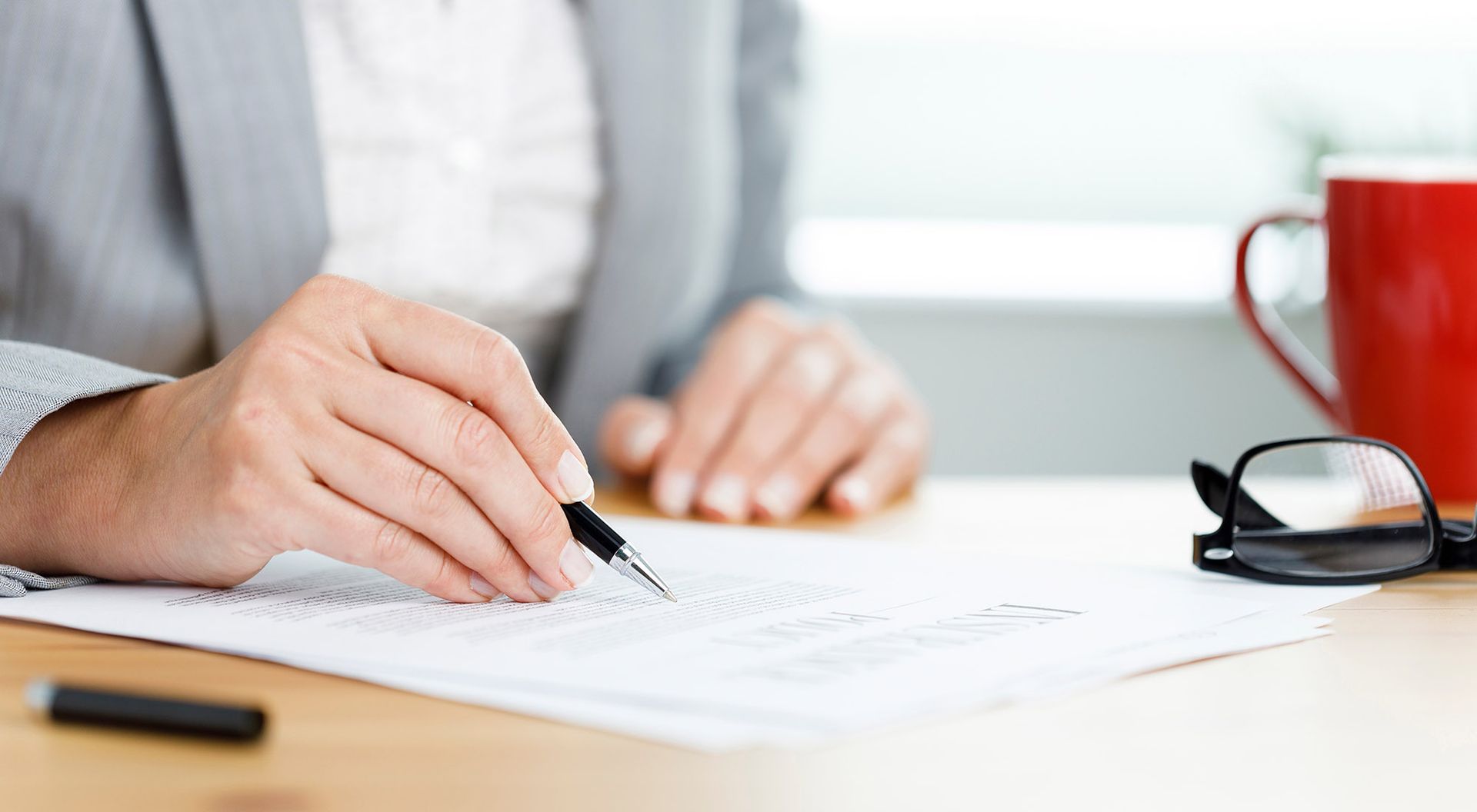 Woman's hand holding a pen, writing on a document at a desk, with glasses and a red mug nearby.