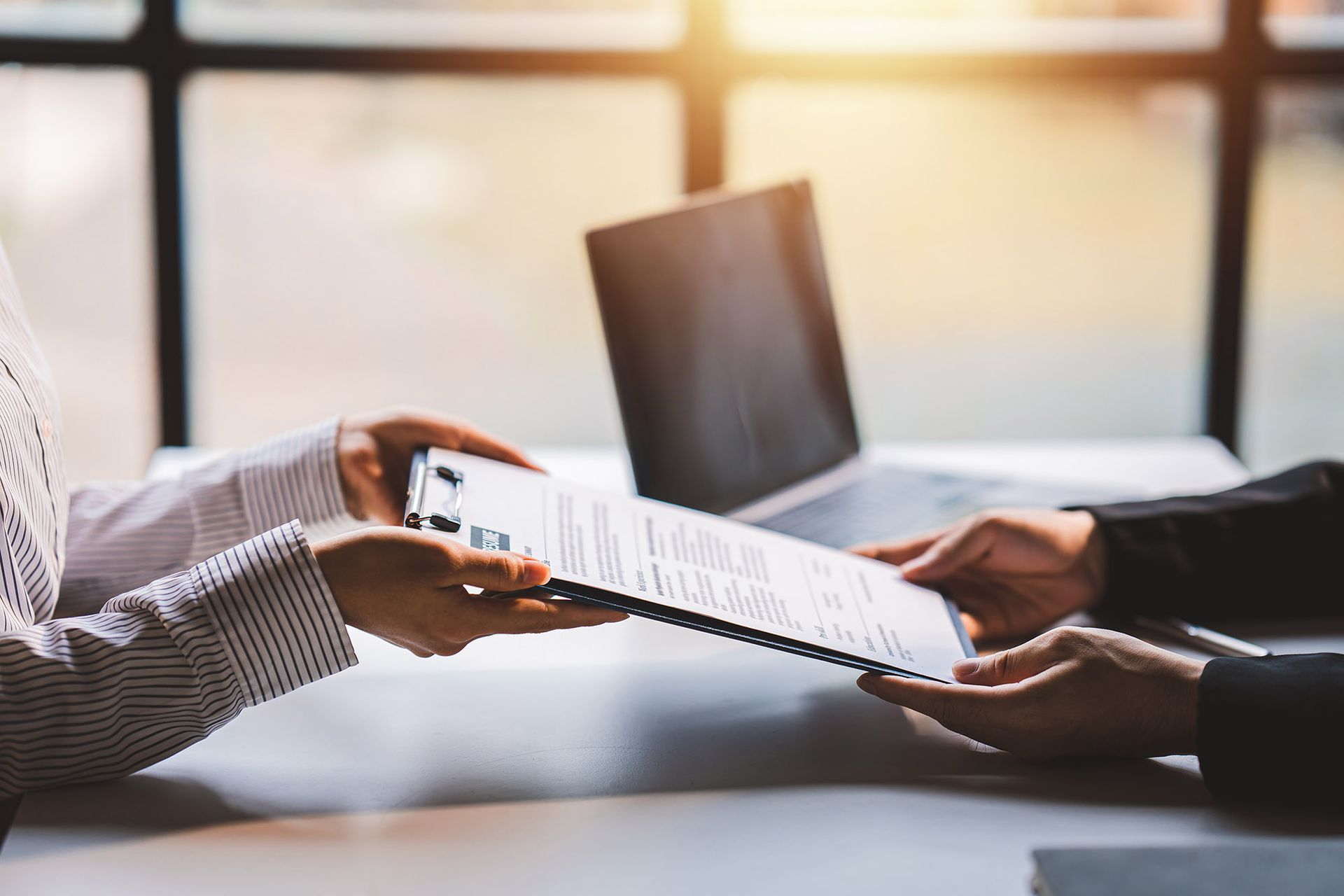 Two people exchanging document on clipboard; laptop in background.