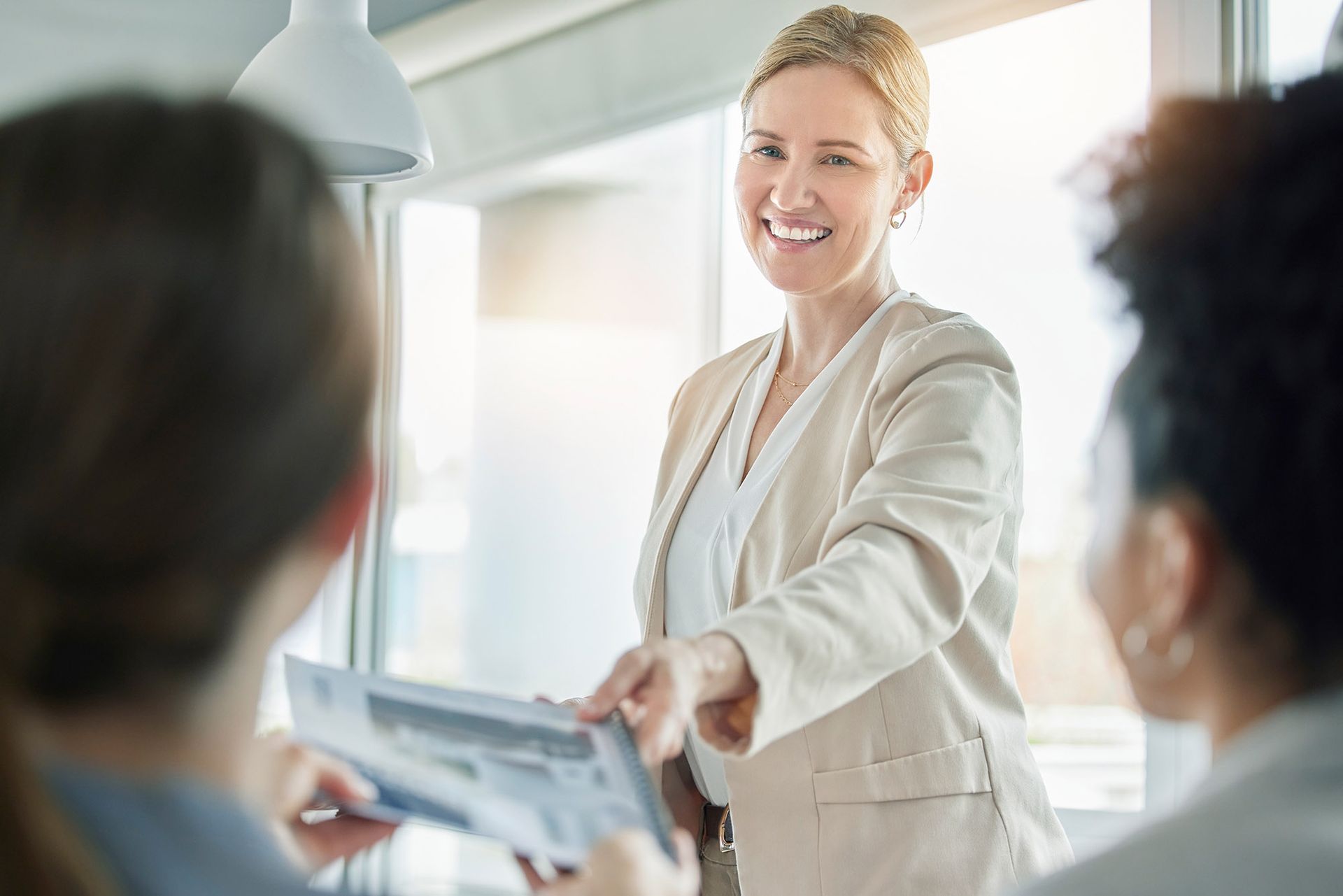 Woman in tan blazer smiles and hands documents to another woman in an office setting.