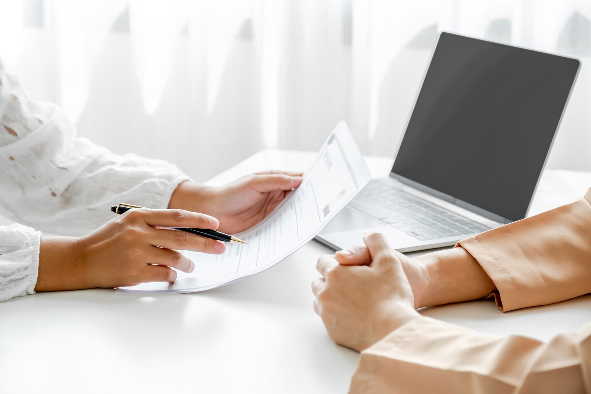 Hands reviewing a document with a pen, next to a laptop on a white desk.