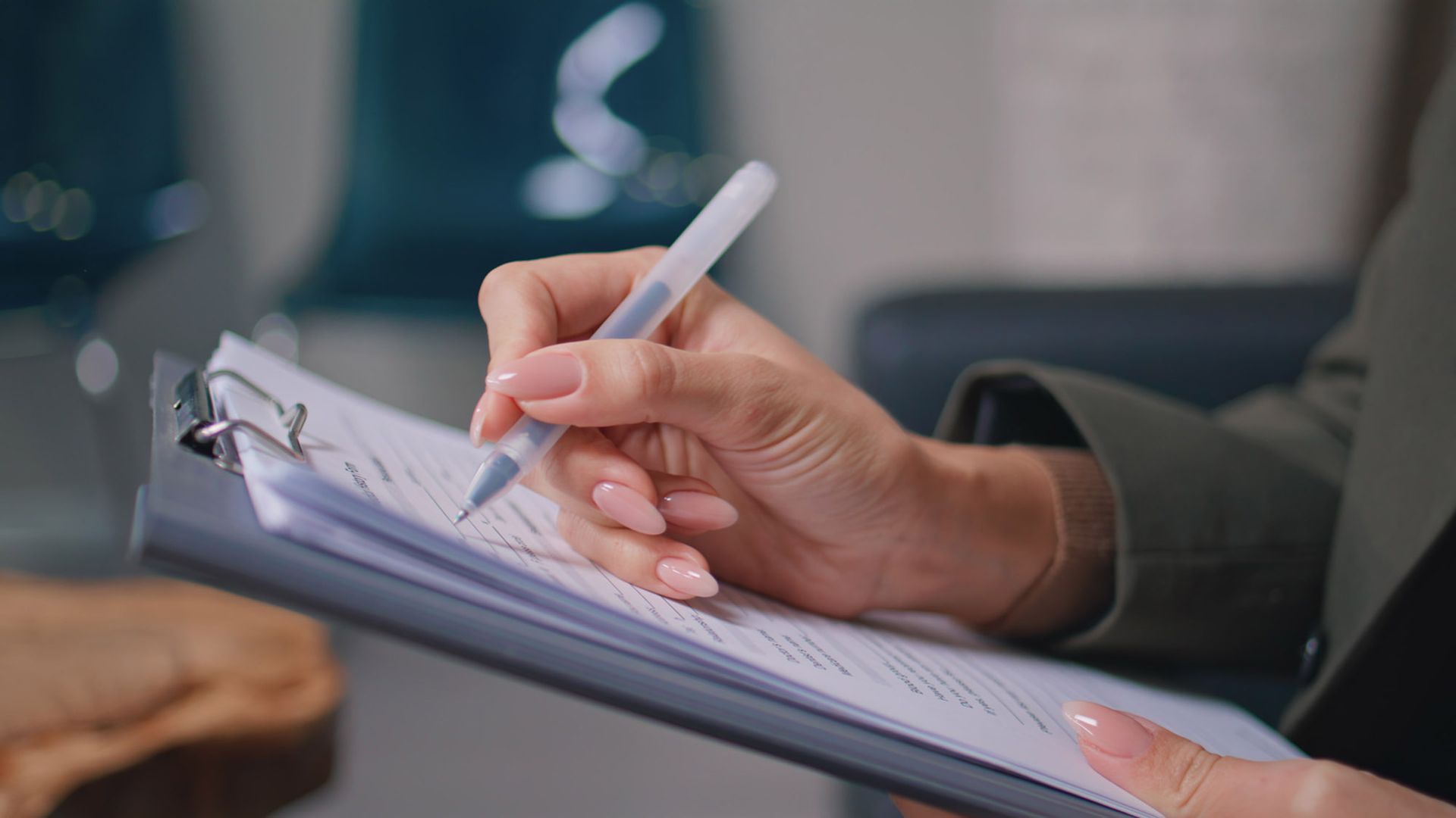 Person's hand writing with a pen on a clipboard holding documents.