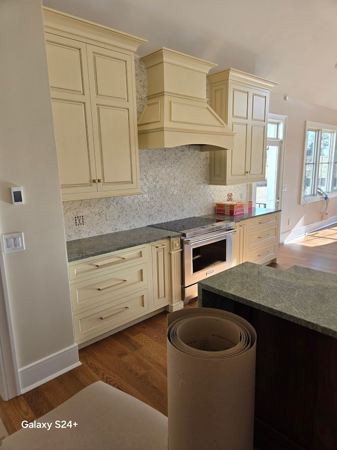 Cream-colored kitchen cabinets with a matching range hood, grey countertops, and a tile backsplash in a bright room.