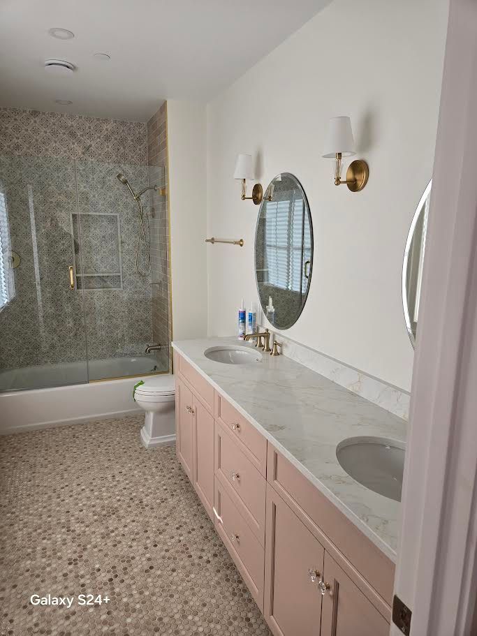 A bright bathroom with pink vanity cabinets, a white marble countertop, dual sinks, oval mirrors, and terrazzo flooring.