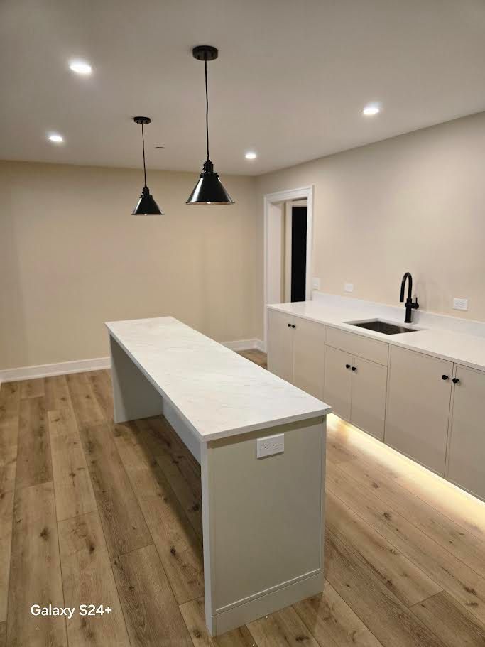 Modern kitchen with a long white island, light cabinets, black faucet, and two black pendant lights over light wood floors.