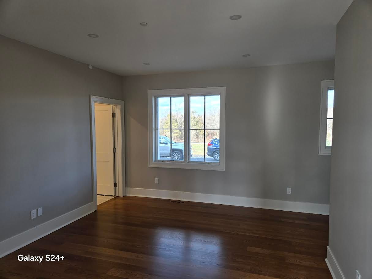 An empty room with light gray walls, dark wood floors, white baseboards, and a large window overlooking a parking area.