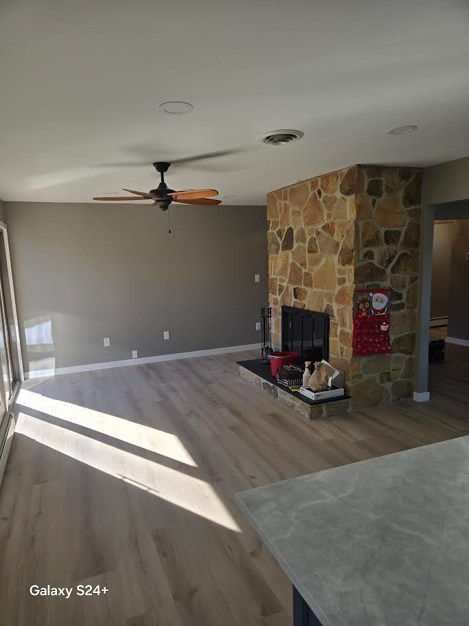 A view of a living room with light wood-style flooring, a stone fireplace, and a ceiling fan.